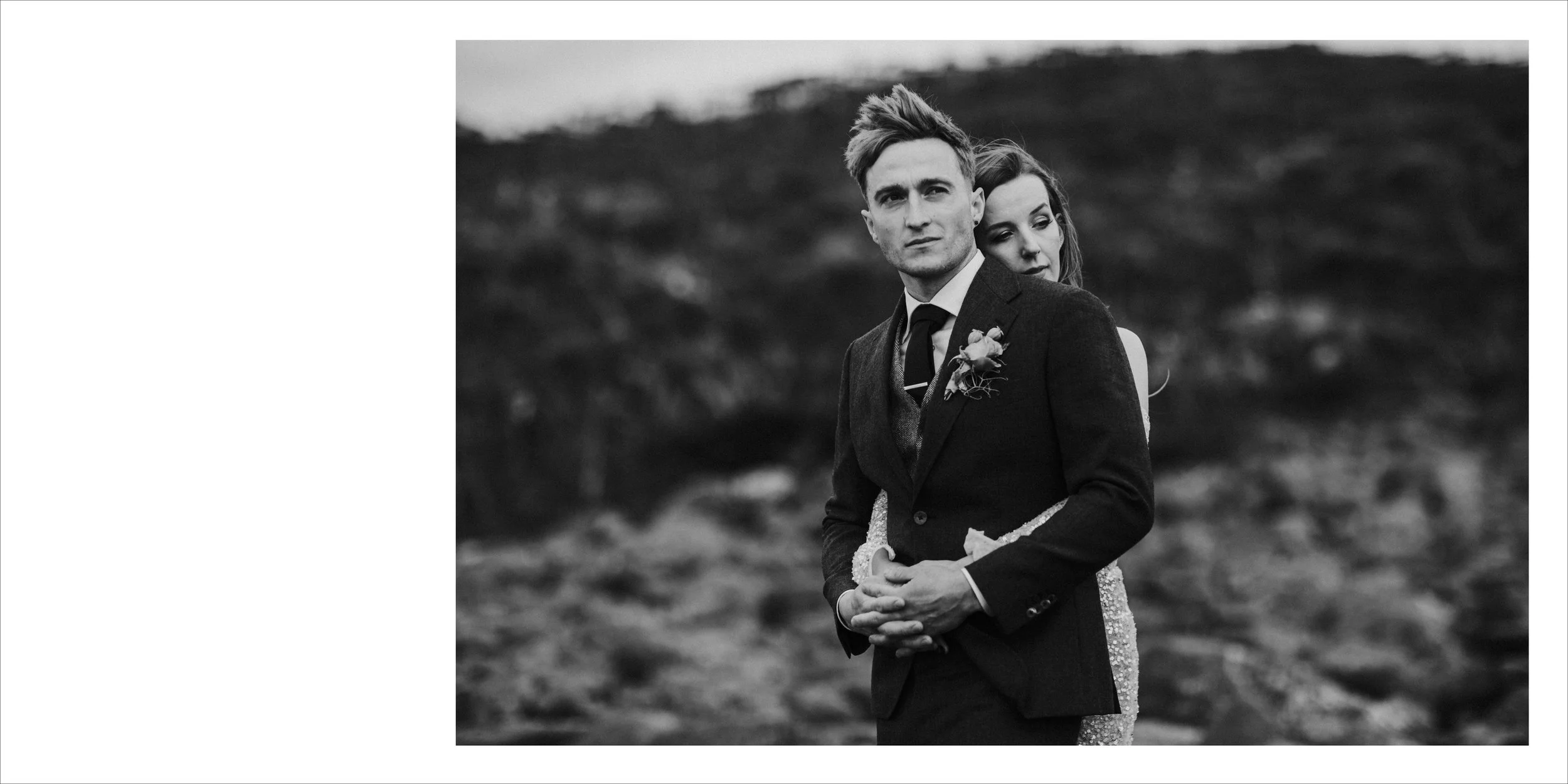Black-and-white photo of a man and woman in a romantic pose outdoors, with the man in a suit and the woman embracing him from behind.