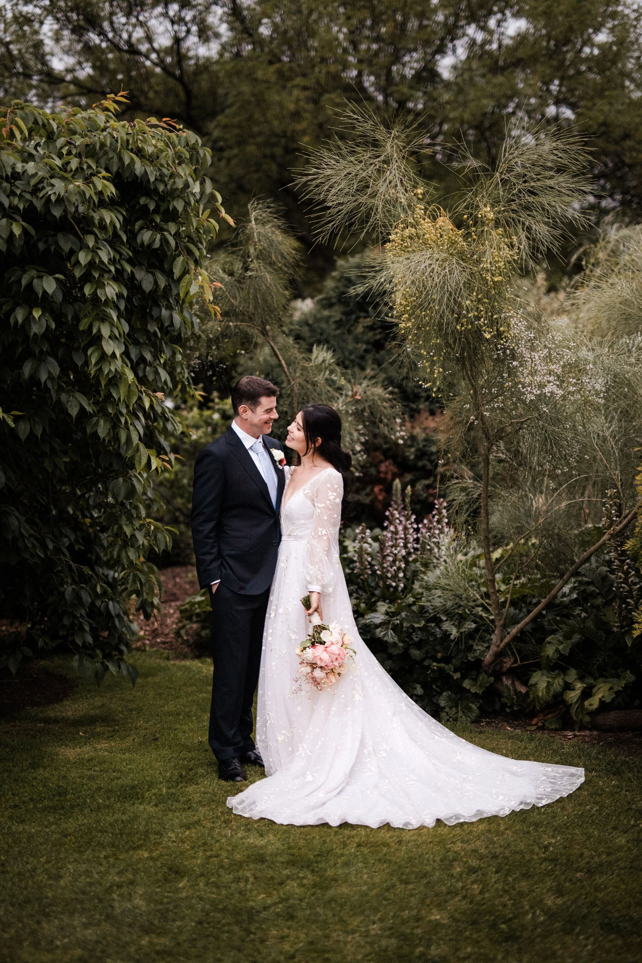 A bride and groom standing close together outdoors, surrounded by lush greenery and flowering plants, on a wedding day.