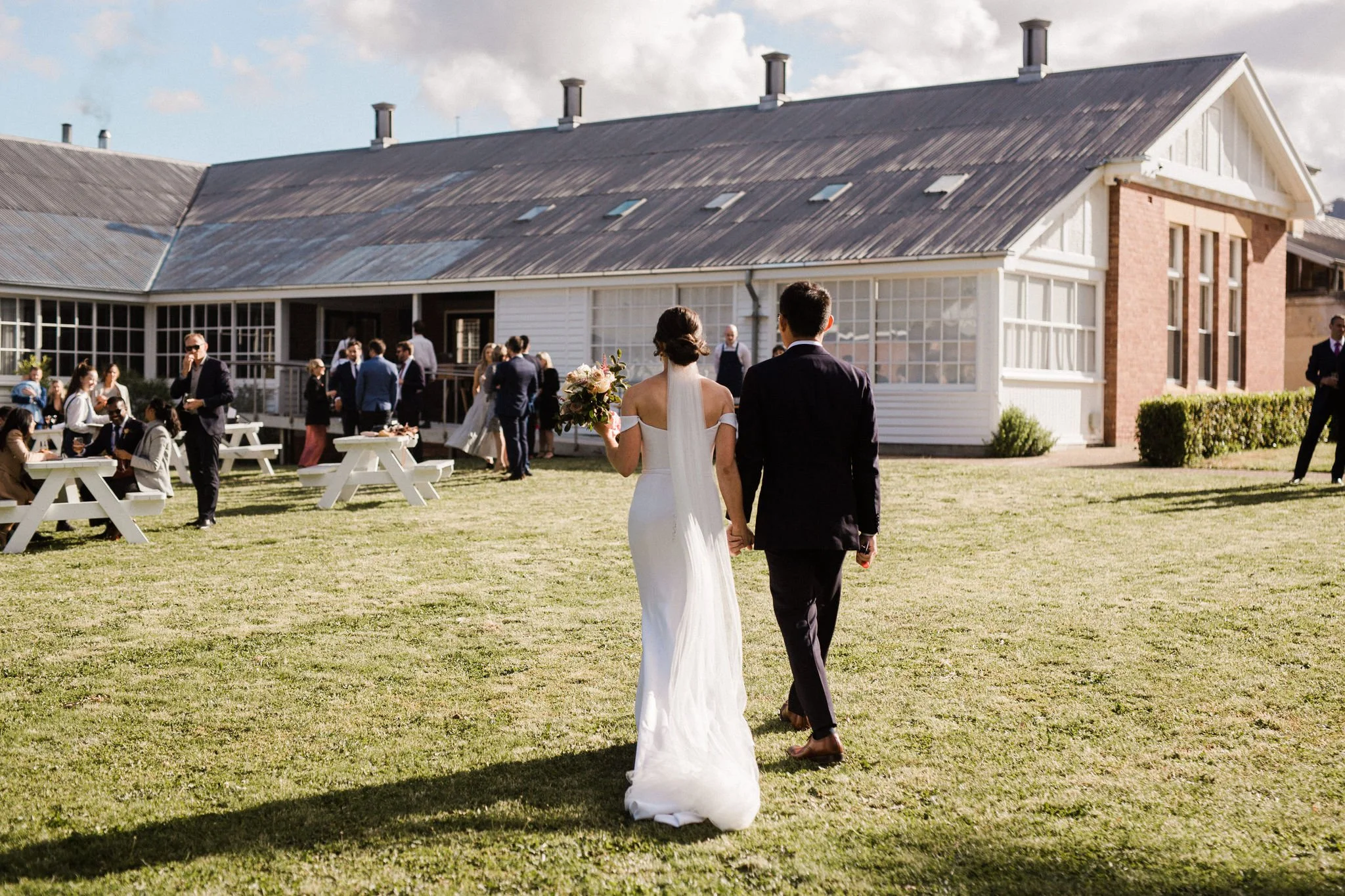 Bride and groom walking hand in hand on a grassy wedding reception outdoor venue with guests in formal attire and a rustic building in the background.