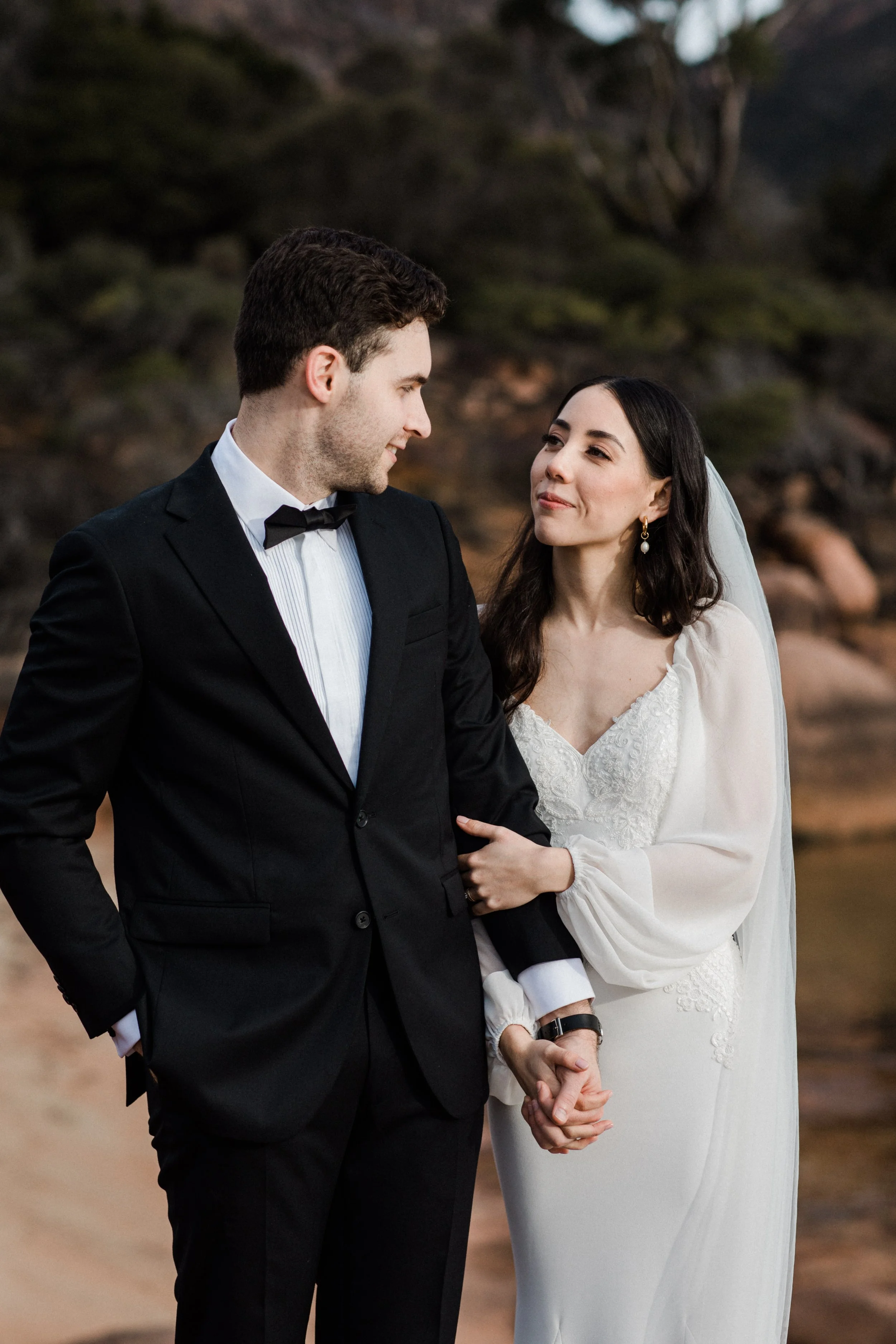 A bride and groom holding hands, gazing at each other during their wedding ceremony outdoors, with rocky terrain and trees in the background.
