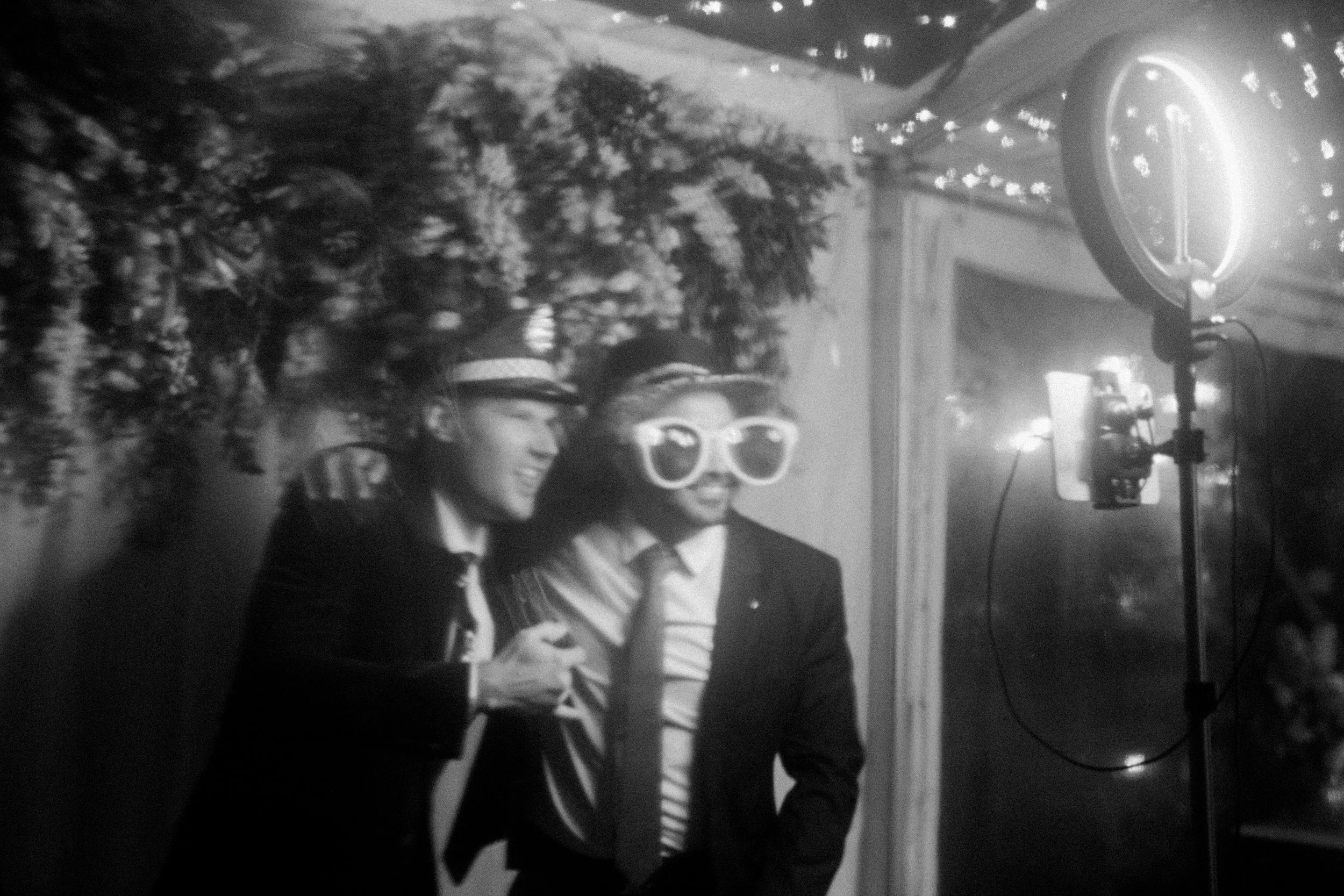 Two people posing for a photo, one woman and one man, both wearing hats and sunglasses. The woman is smiling, and the man has a serious expression. They are in front of a floral backdrop, with photo lighting equipment visible on the right.