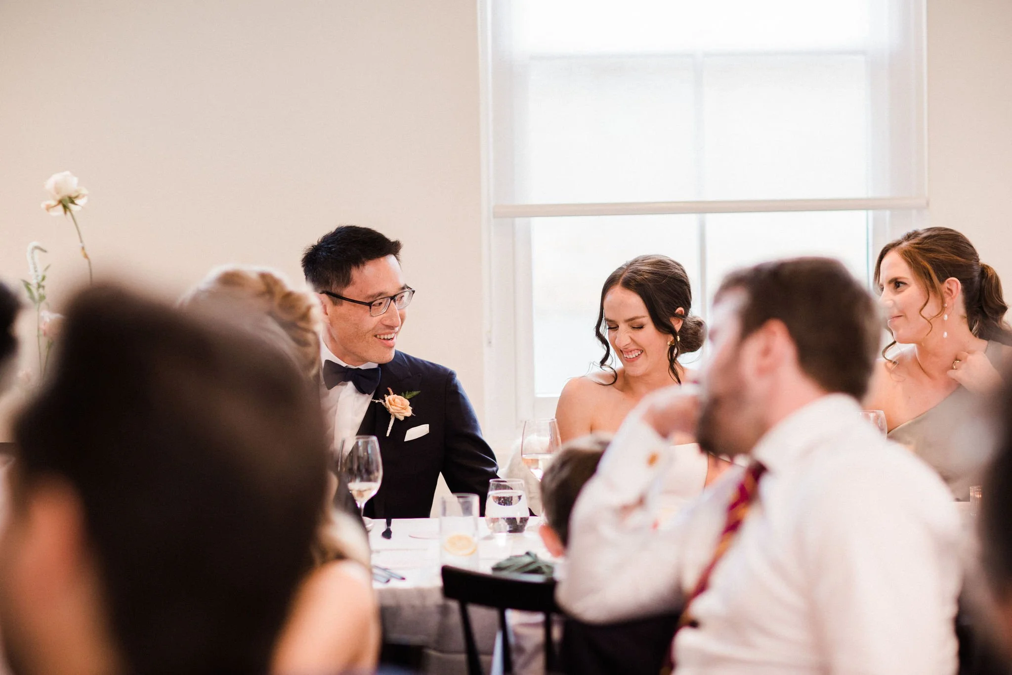 Group of people at a wedding reception sitting around a table, smiling and laughing, with a bride and groom in the center.