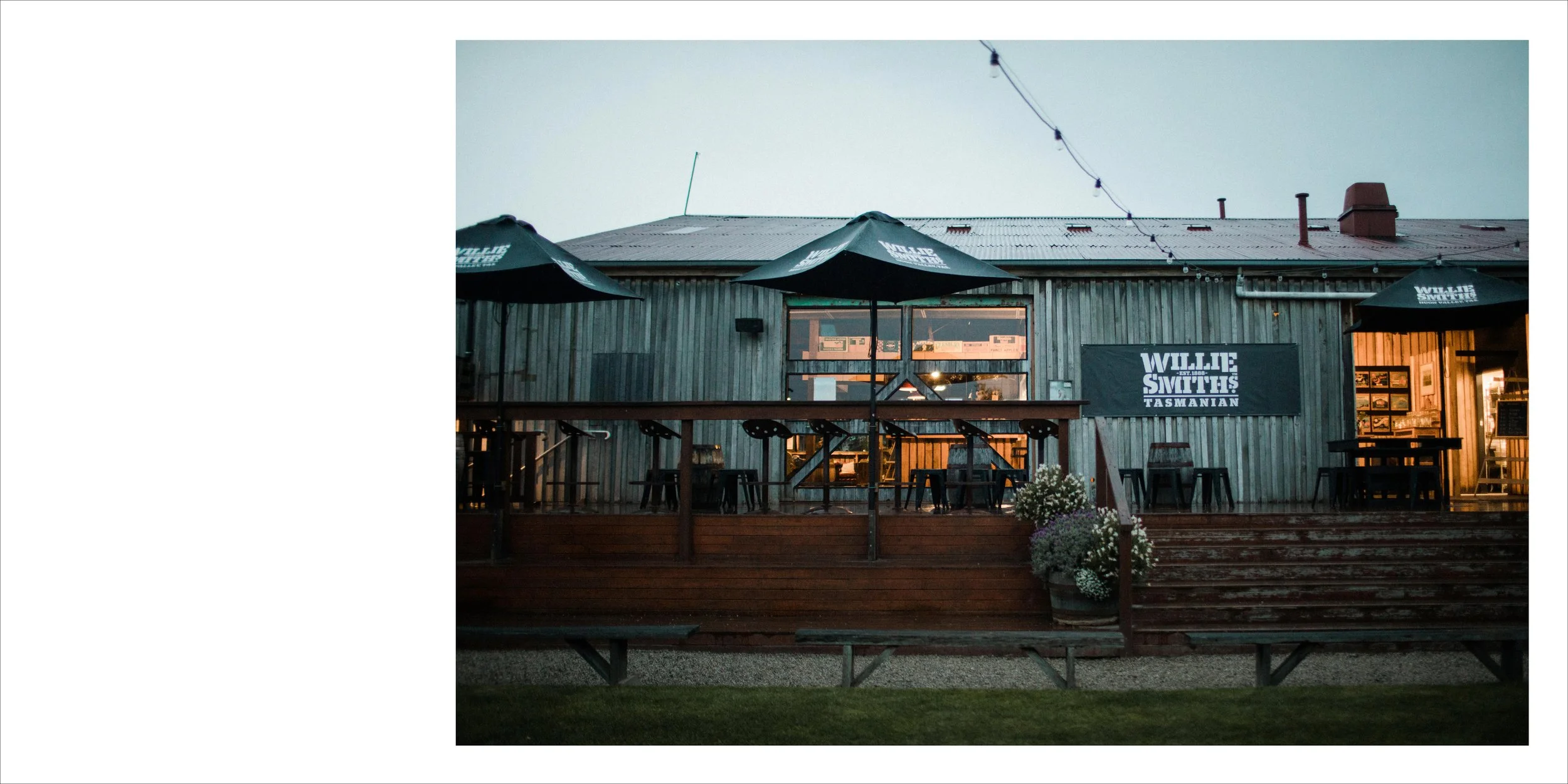 Exterior of a rustic Tasmanian restaurant called Willie Smith's with outdoor seating and umbrellas, illuminated warmly inside during dusk.