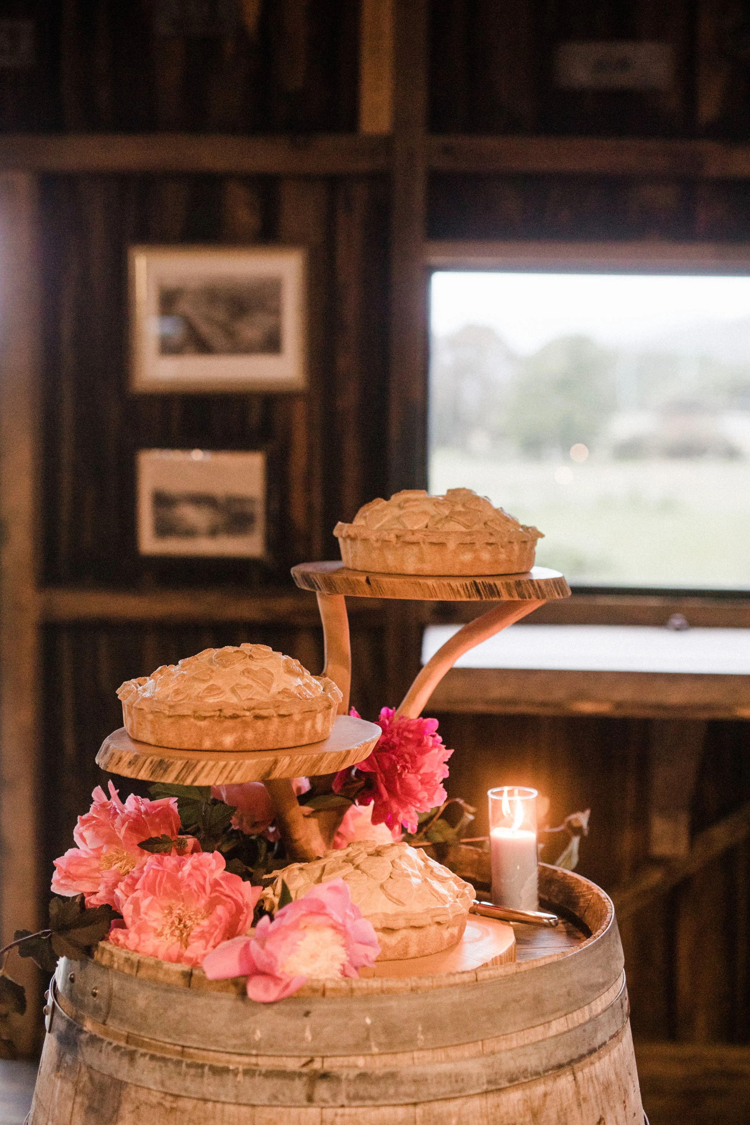Pie display on a wooden barrel decorated with pink flowers and a candle, inside a rustic room with wood-paneled walls and framed photos.