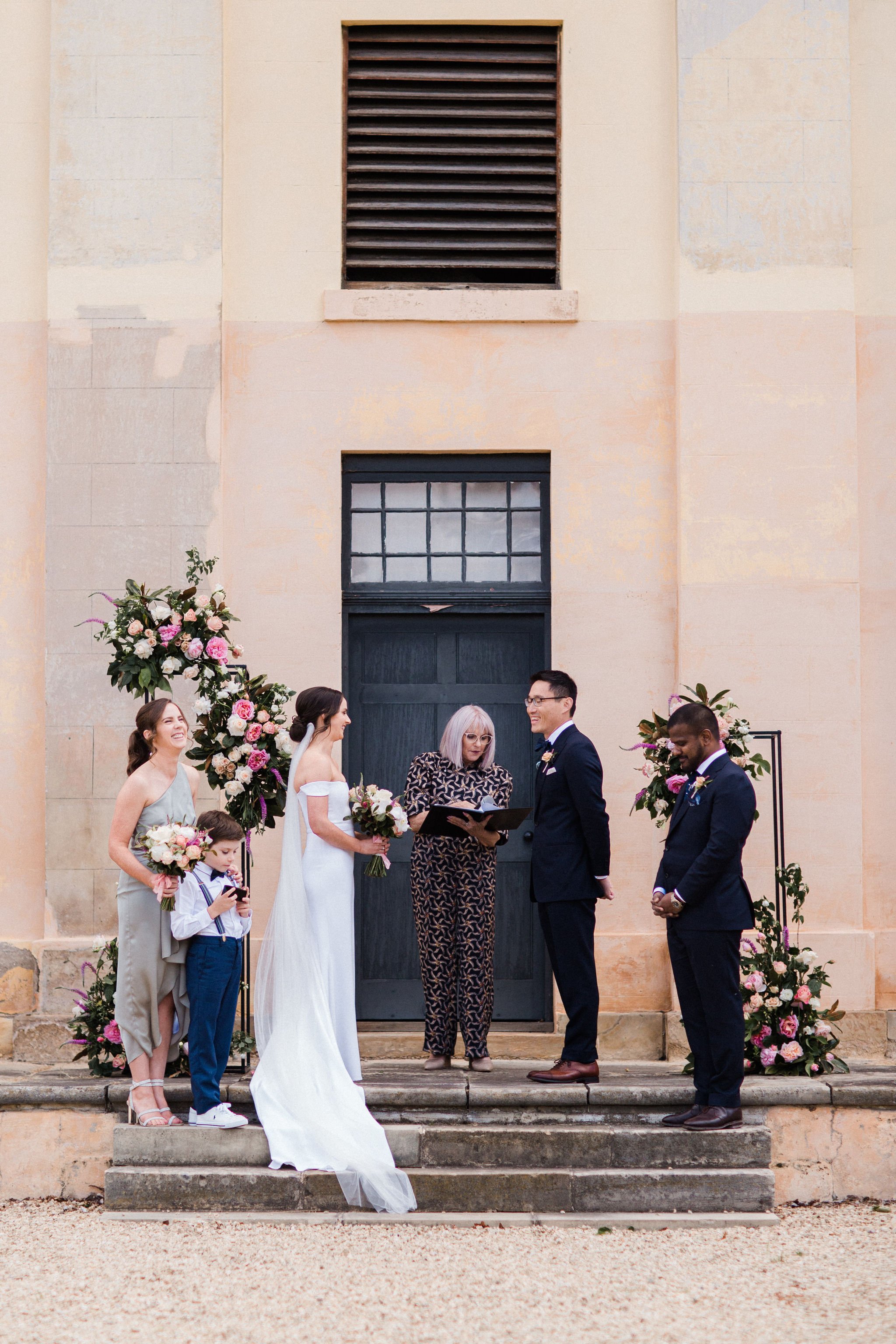 A wedding ceremony taking place outdoors in front of a stone building with a large dark door and high window above it. The bride and groom are standing in front of an officiant, across from a bridesmaid, a child, and an usher or groomsman. There are 