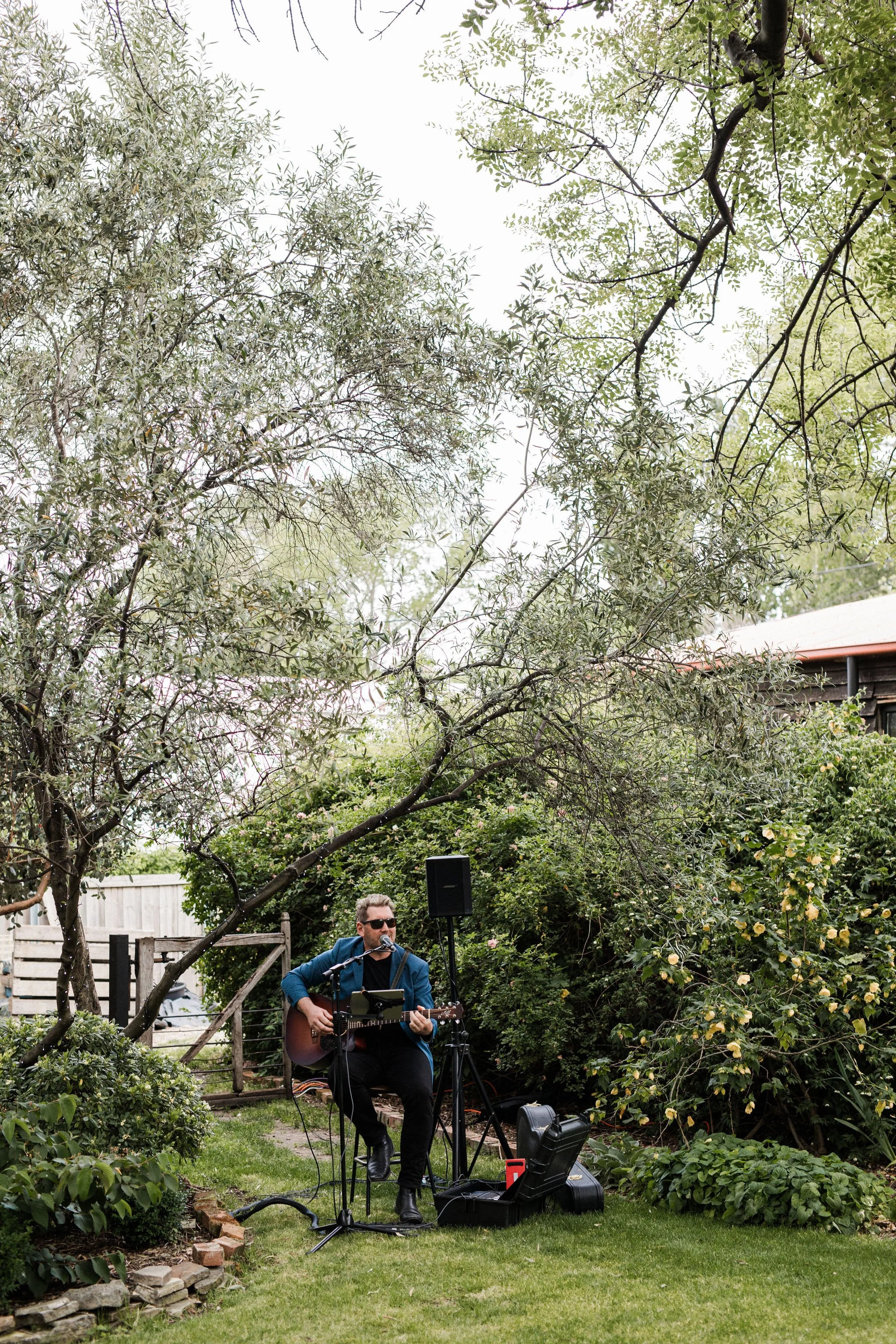 A man with sunglasses, blue jacket, and black pants playing an acoustic guitar and singing into a microphone in a garden, surrounded by trees and bushes.