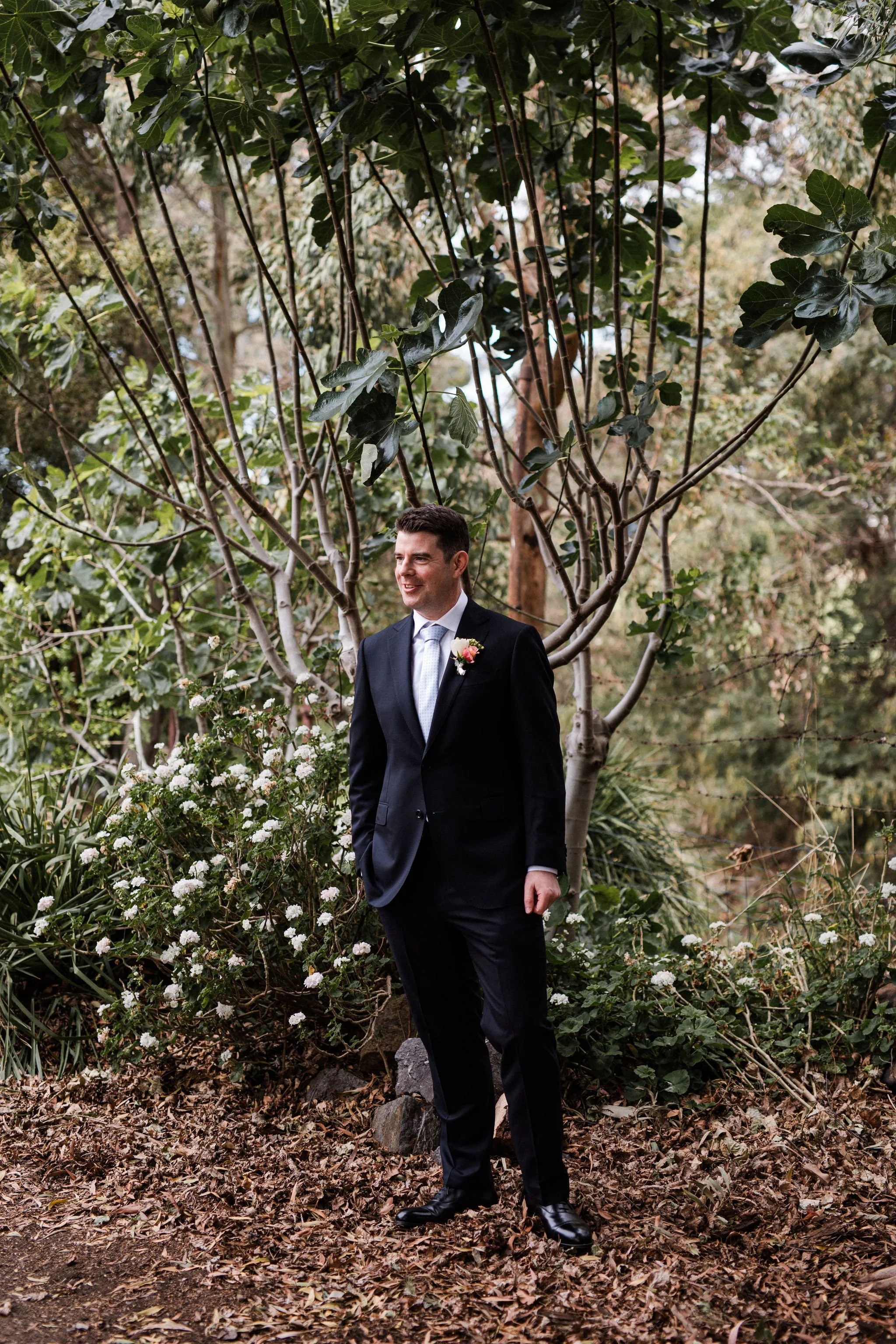 A man dressed in a dark suit with a boutonniere stands outdoors among trees and plants, smiling.