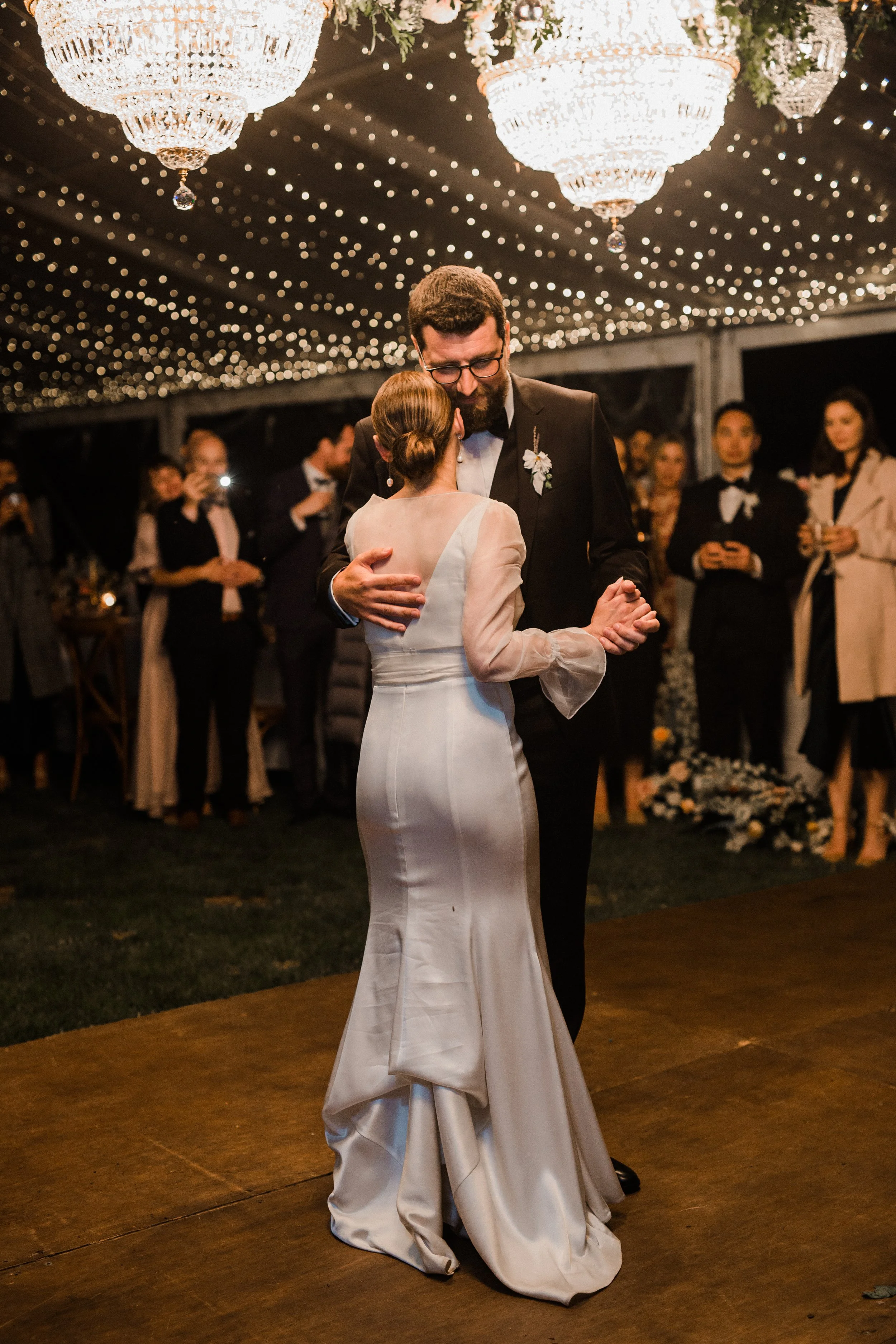 A bride and groom sharing a dance at their wedding reception, under romantic chandeliers and string lights, with guests watching in the background.