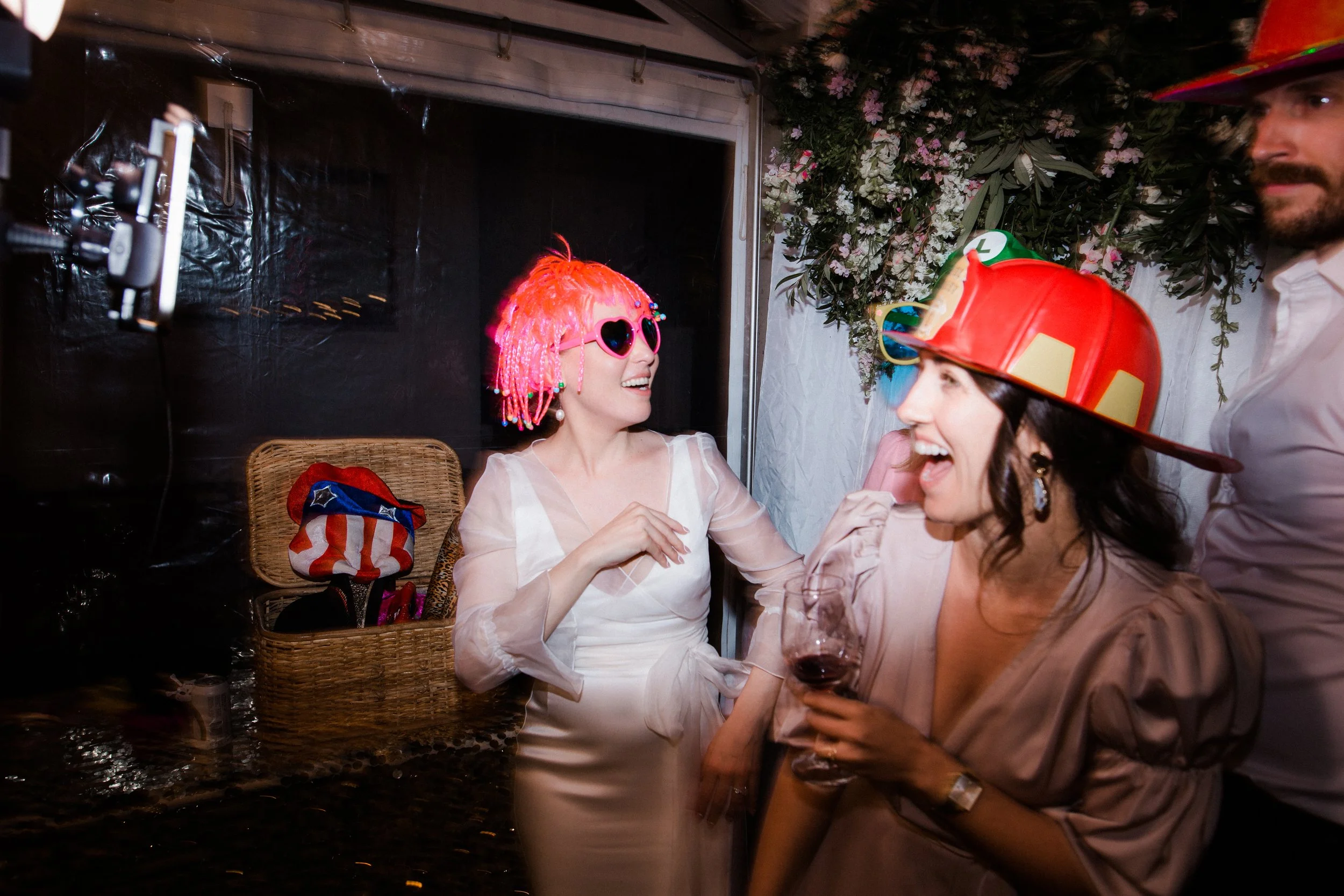 Three women celebrating at a party, wearing colorful hats and glasses, with one woman holding a glass of wine, all smiling and laughing.