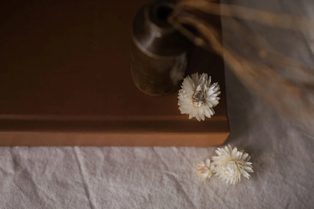 Close-up of a small white flower resting on a brown surface next to a ceramic vase, with the background showing a textured light-colored cloth.