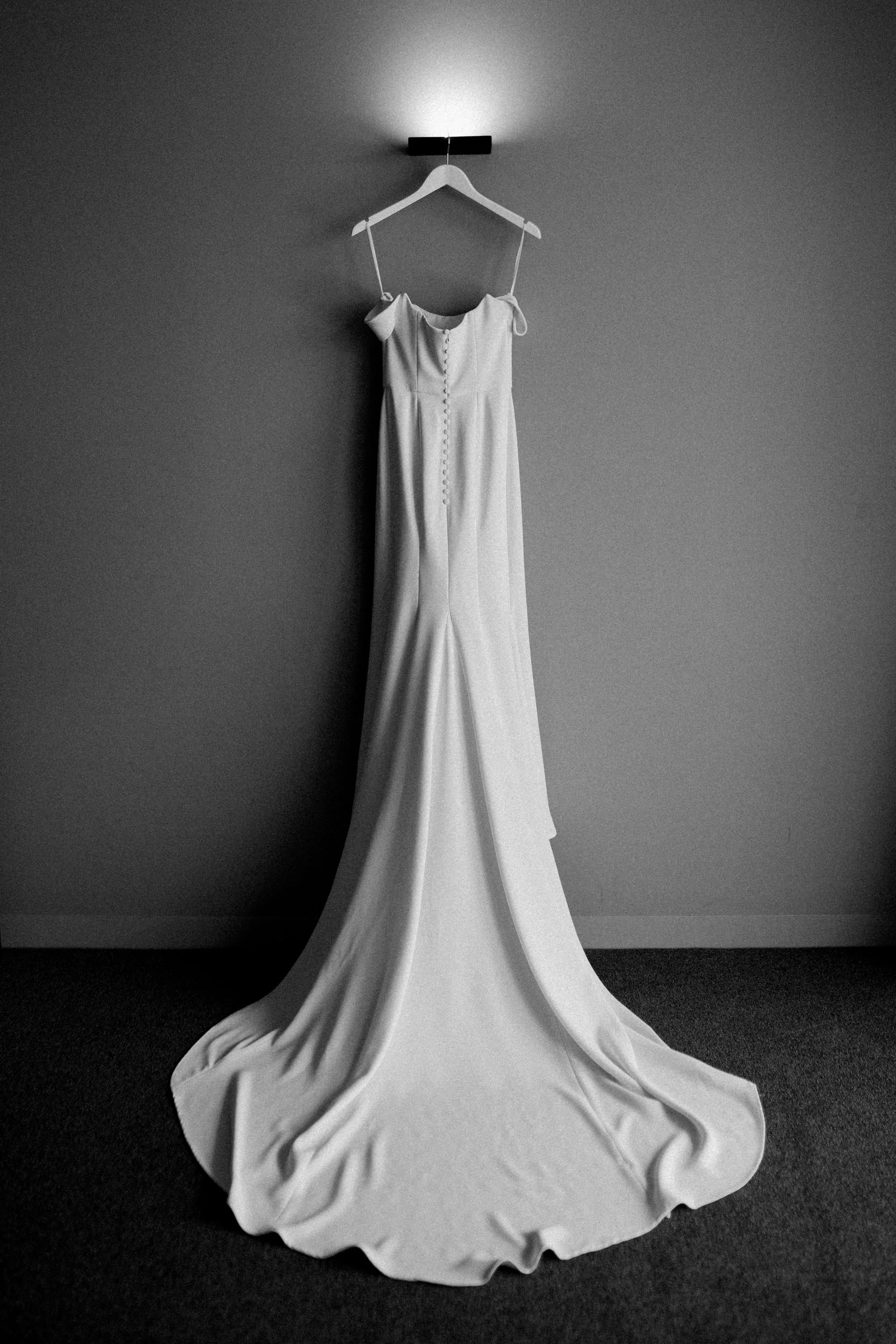 A white wedding dress hanging on a white hanger against a plain wall, with the dress pooling on the dark floor.