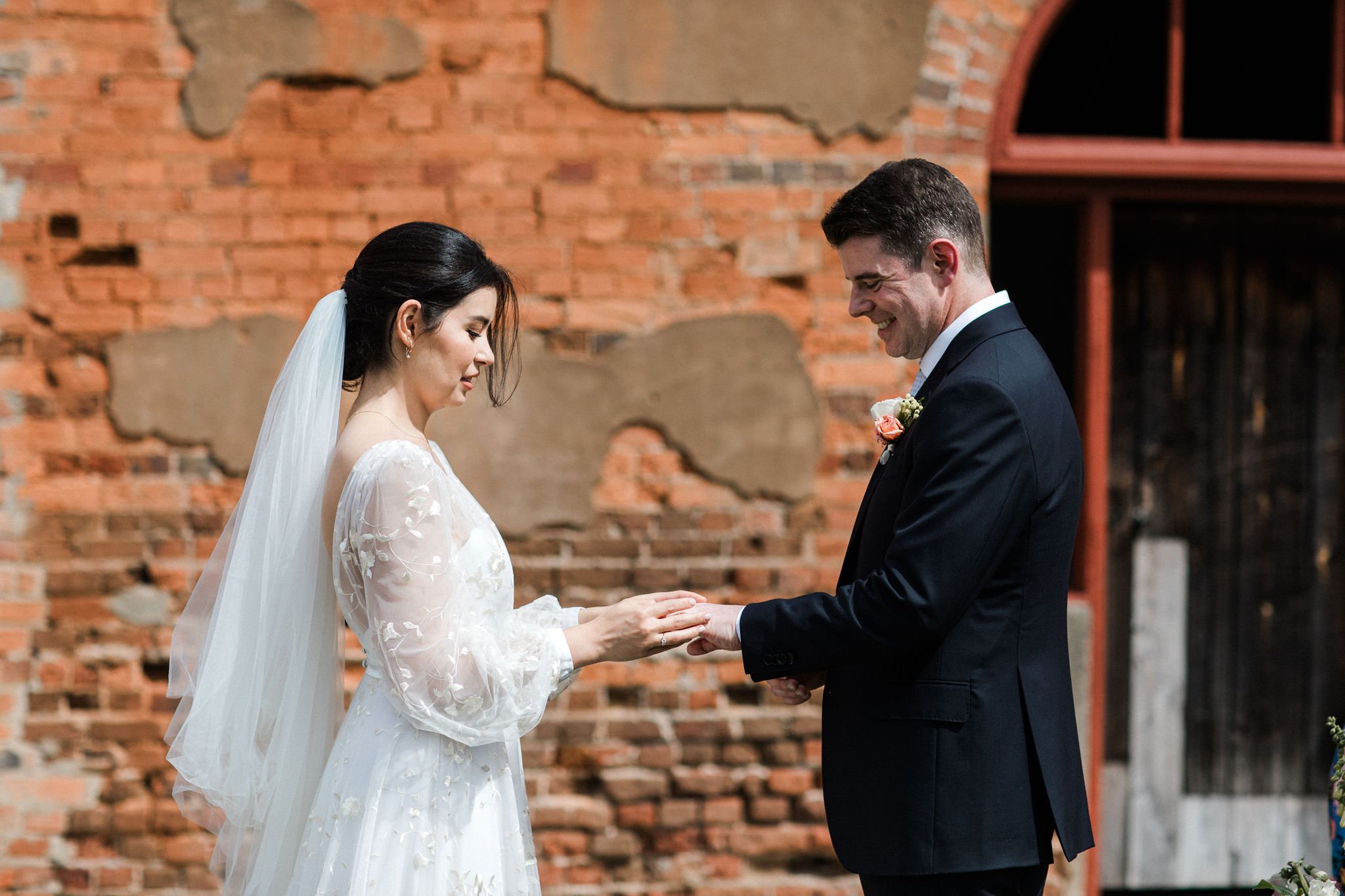 A bride and groom exchanging rings during a wedding ceremony outside against a rustic brick wall.