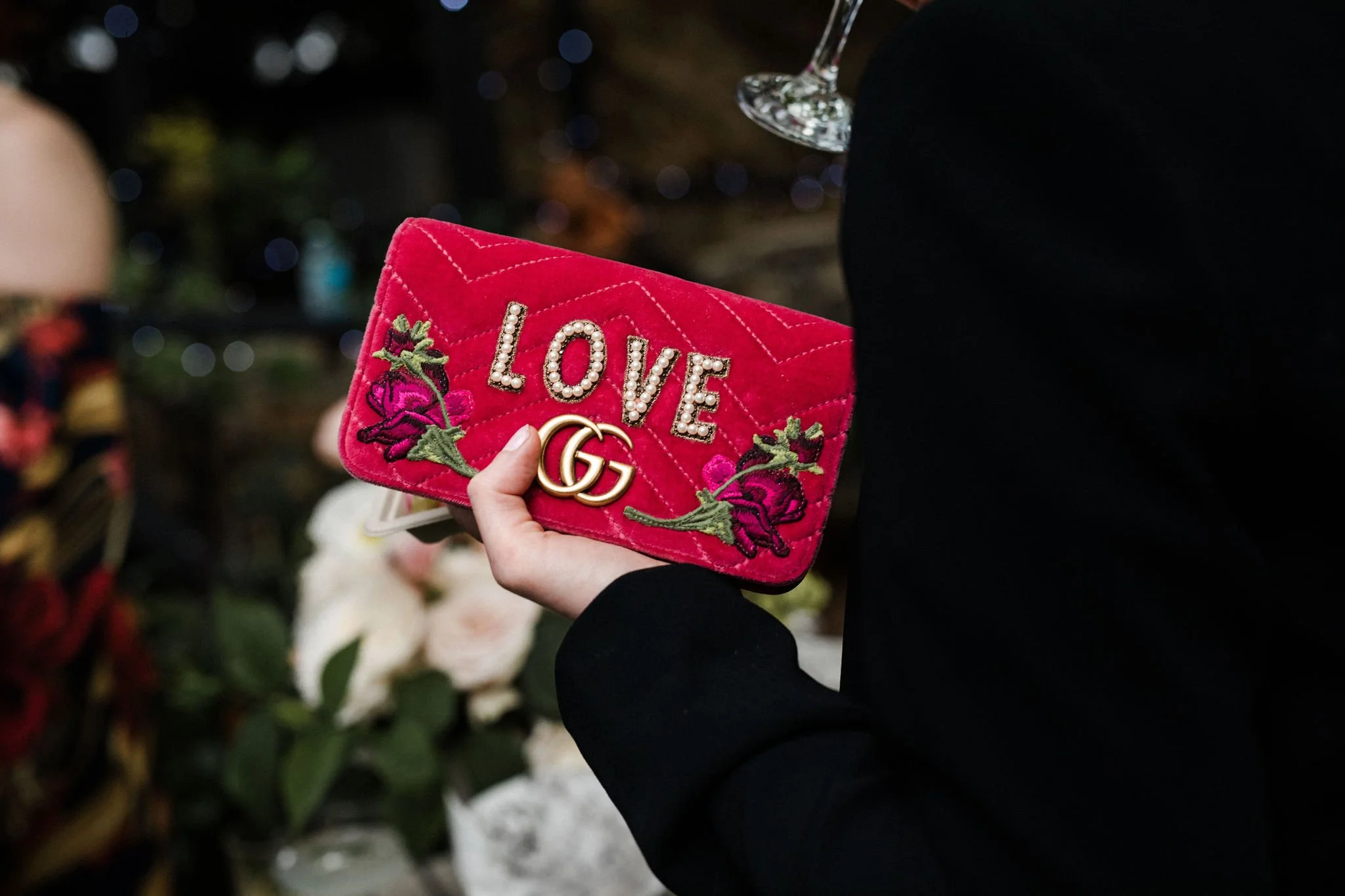 Person holding a pink velvet purse with the word 'LOVE' decorated with pearls, surrounded by embroidered purple flowers, and gold buckles, with a glass of wine in the background.