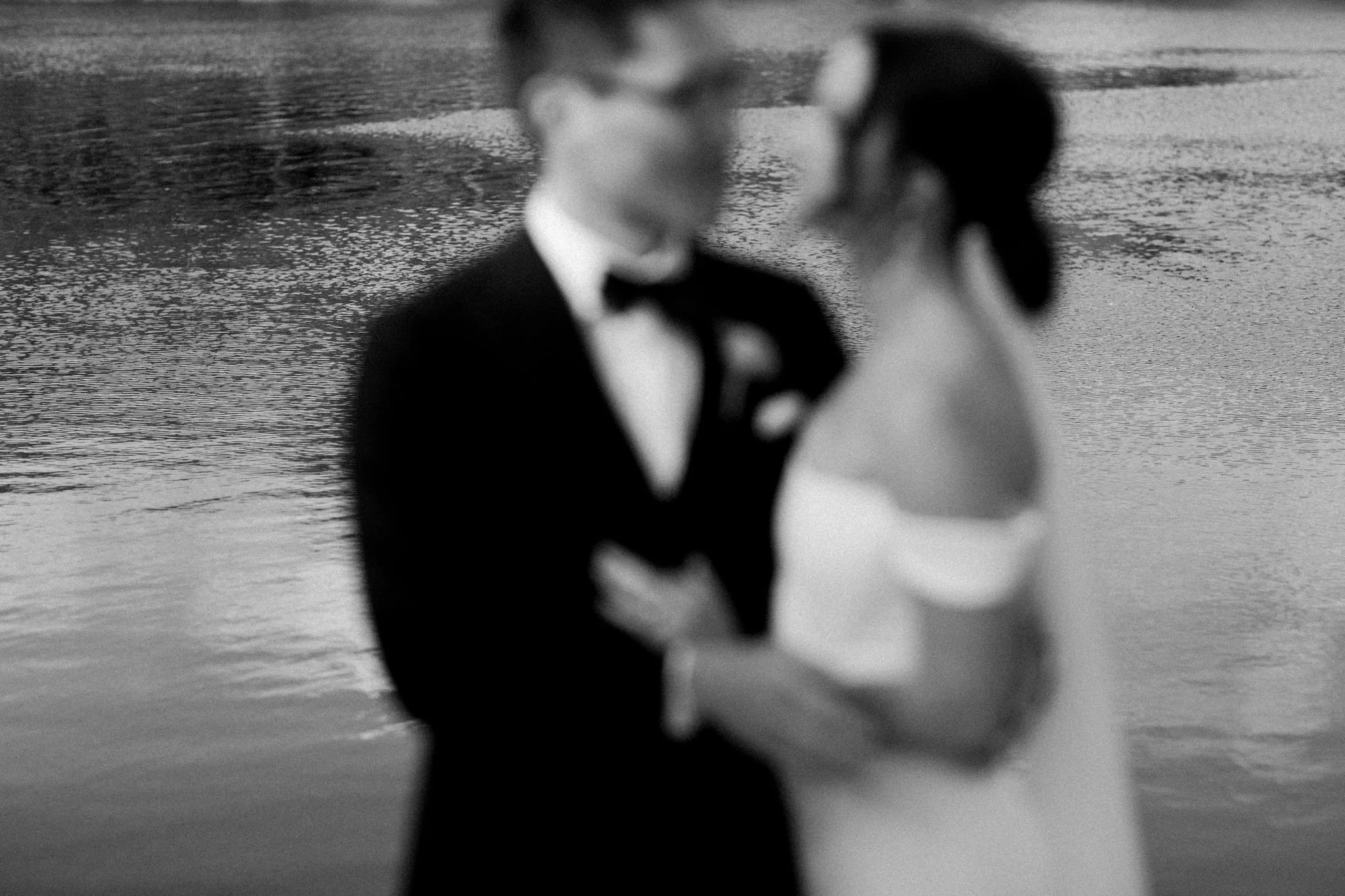 Black and white photo of a bride and groom holding hands near water, blurred effect.