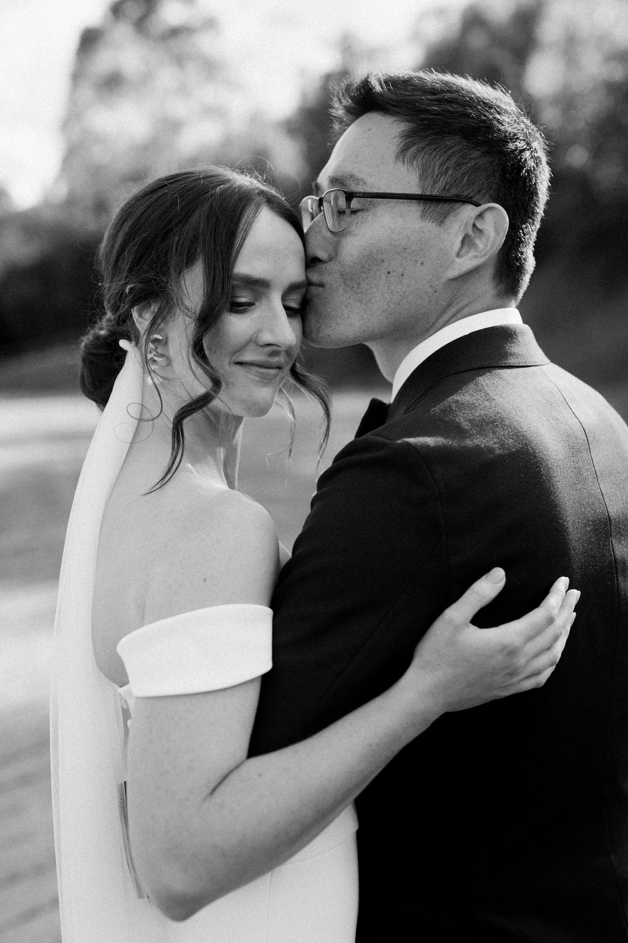 A black-and-white photo of a bride and groom embracing outdoors. The groom is kissing the bride on her forehead, with her eyes closed and a gentle smile. She is wearing an off-shoulder wedding dress and a veil, while he is in a suit with spectacles.