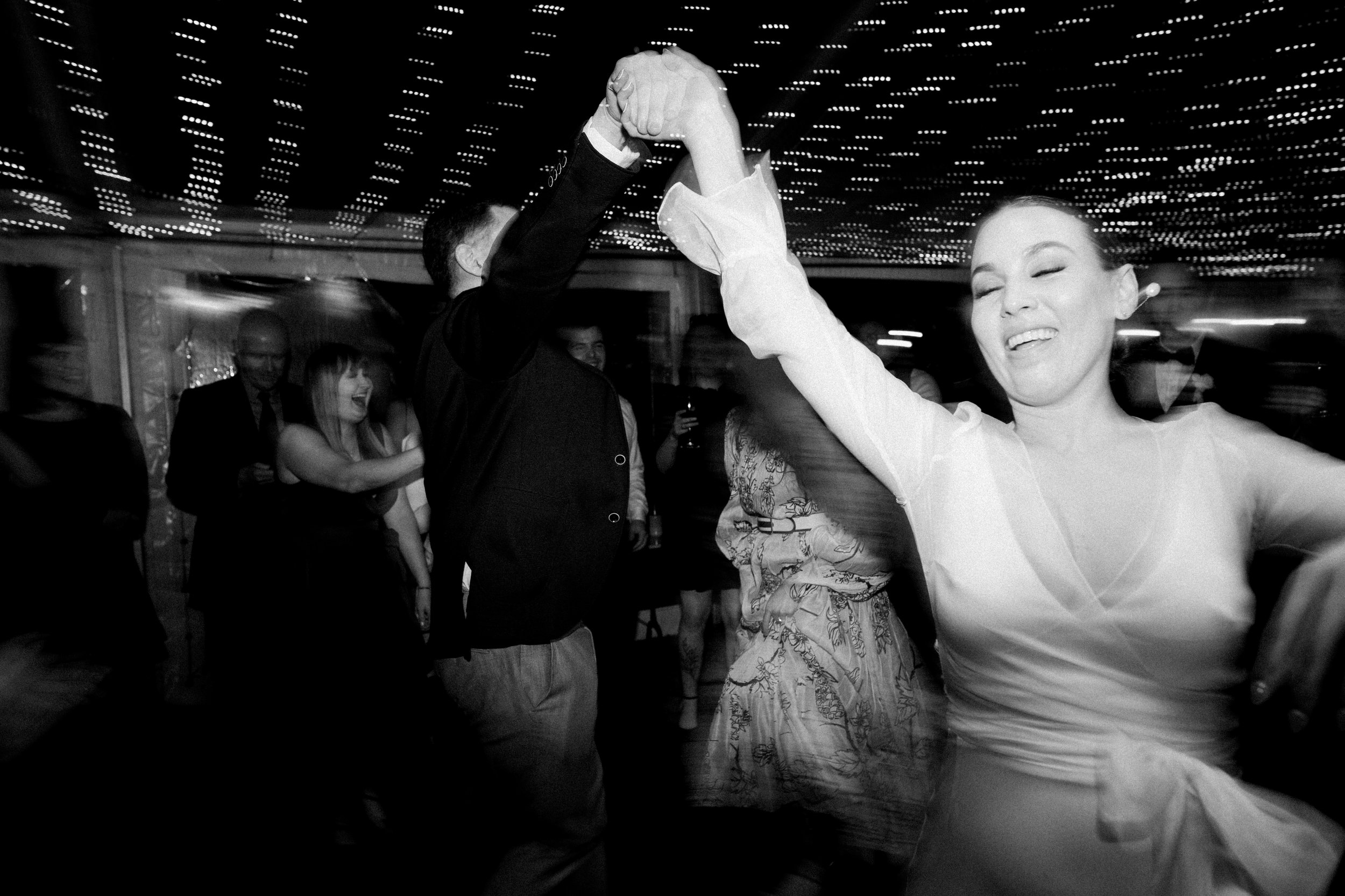 People dancing at a celebration, with a woman smiling and a man holding her hand in a dance. The ceiling is decorated with string lights, creating a festive atmosphere.