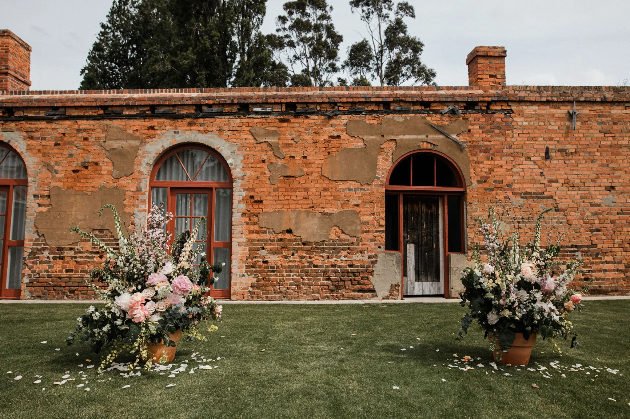 A brick building with three arched windows, some with lace curtains, and a door, surrounded by landscaped grass with two large flower arrangements in terracotta pots with pink and white flowers and petals scattered on the ground in front.