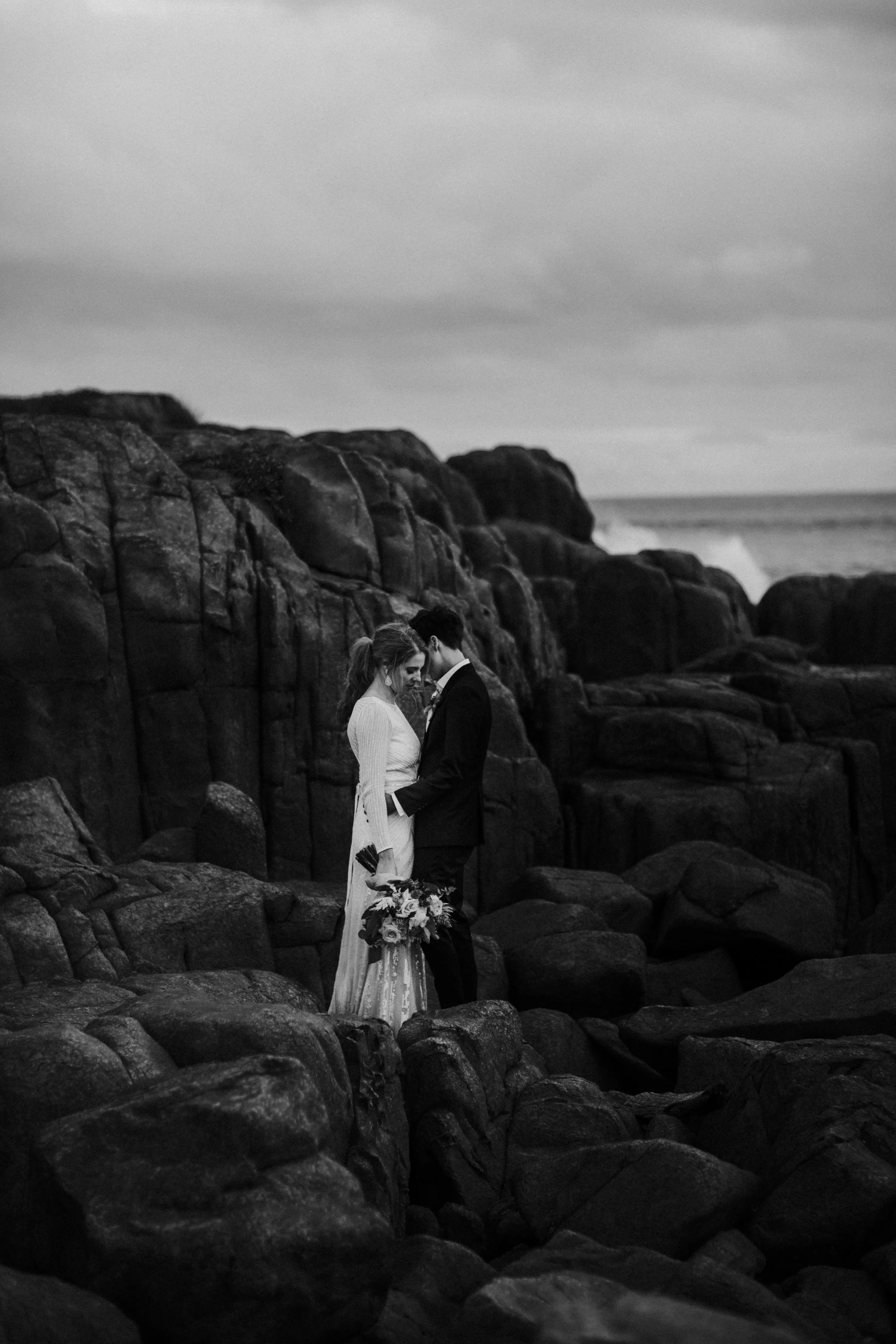 A black and white photo of a bride and groom standing close on a rocky shoreline, holding hands, with the bride holding a bouquet of flowers, and ocean waves in the background.
