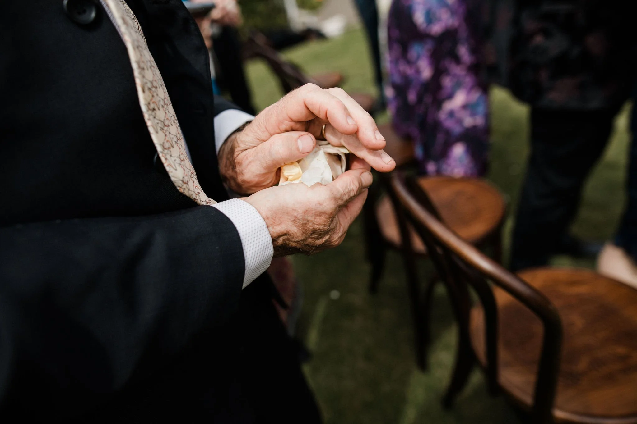 Close-up of an elderly man holding a small object, possibly a wrapped gift or paper, with other people and chairs in the background at an outdoor event.