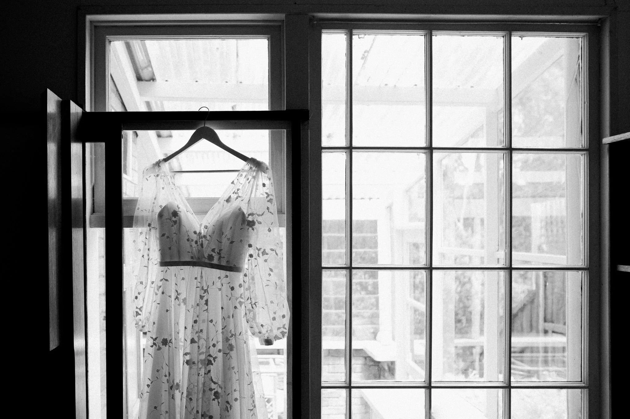 A wedding dress with floral pattern hanging on a black hanger in front of a large window with multiple panes.