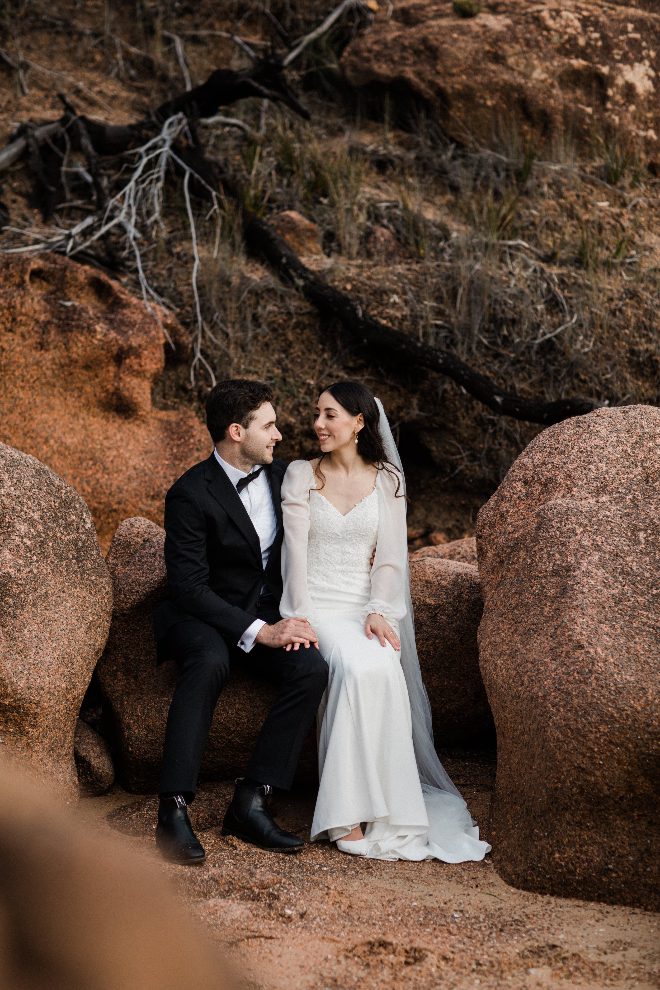 A bride and groom sitting on rocks in a desert landscape, smiling and looking at each other, with large reddish-brown rocks and sparse vegetation in the background.