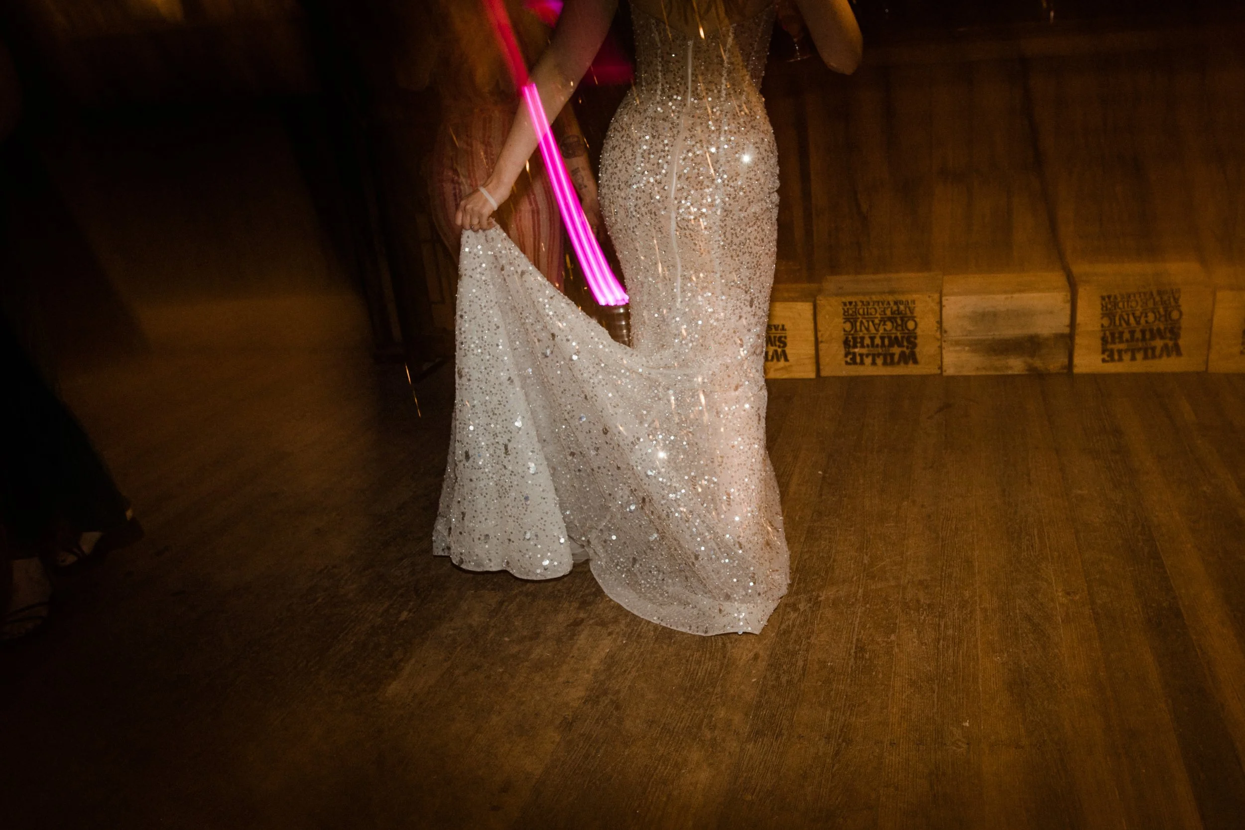 Person in a sparkling, glittery gown dancing with a glow stick in a dimly lit wooden venue.