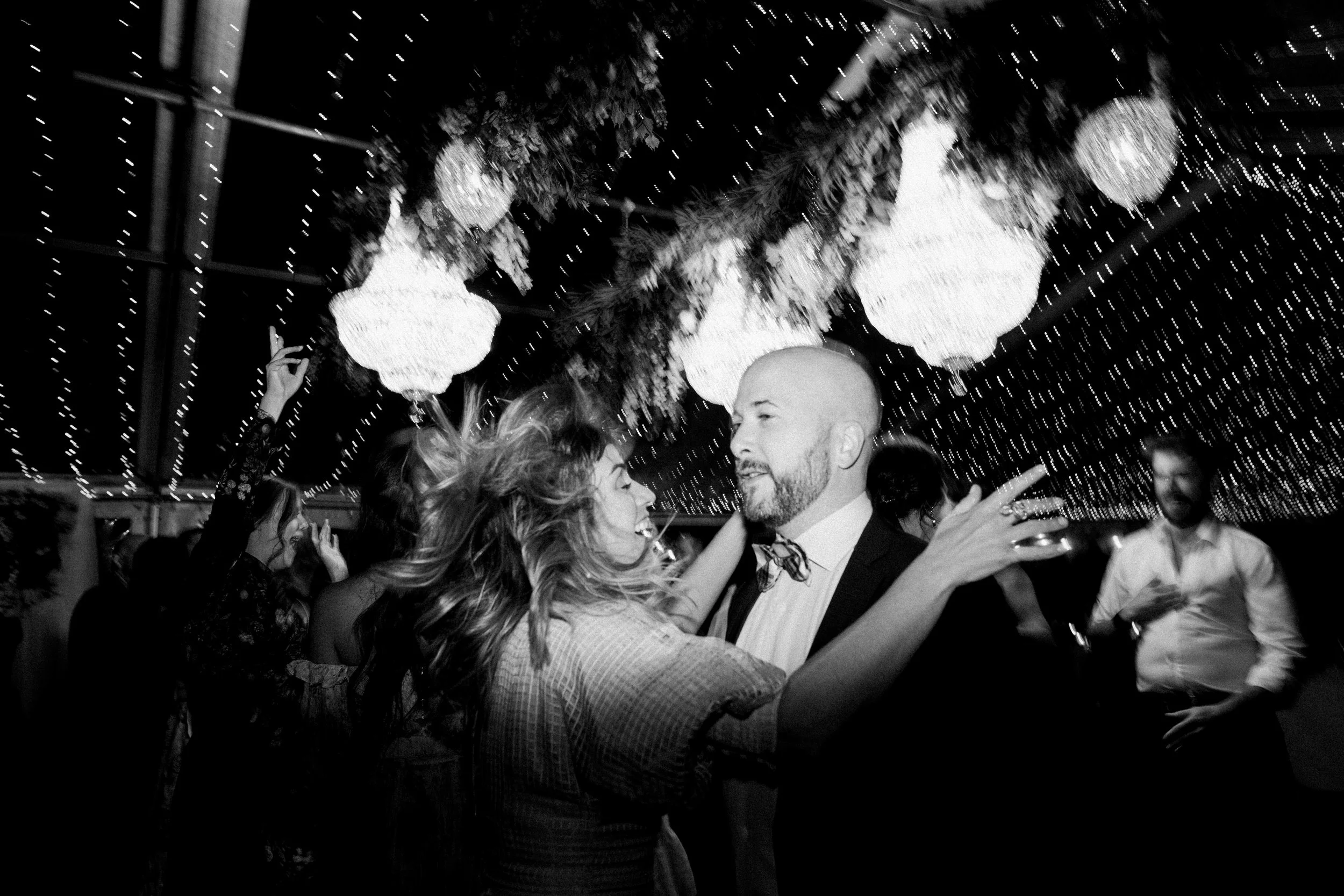 Couple dancing closely under hanging chandeliers and string lights at a party.