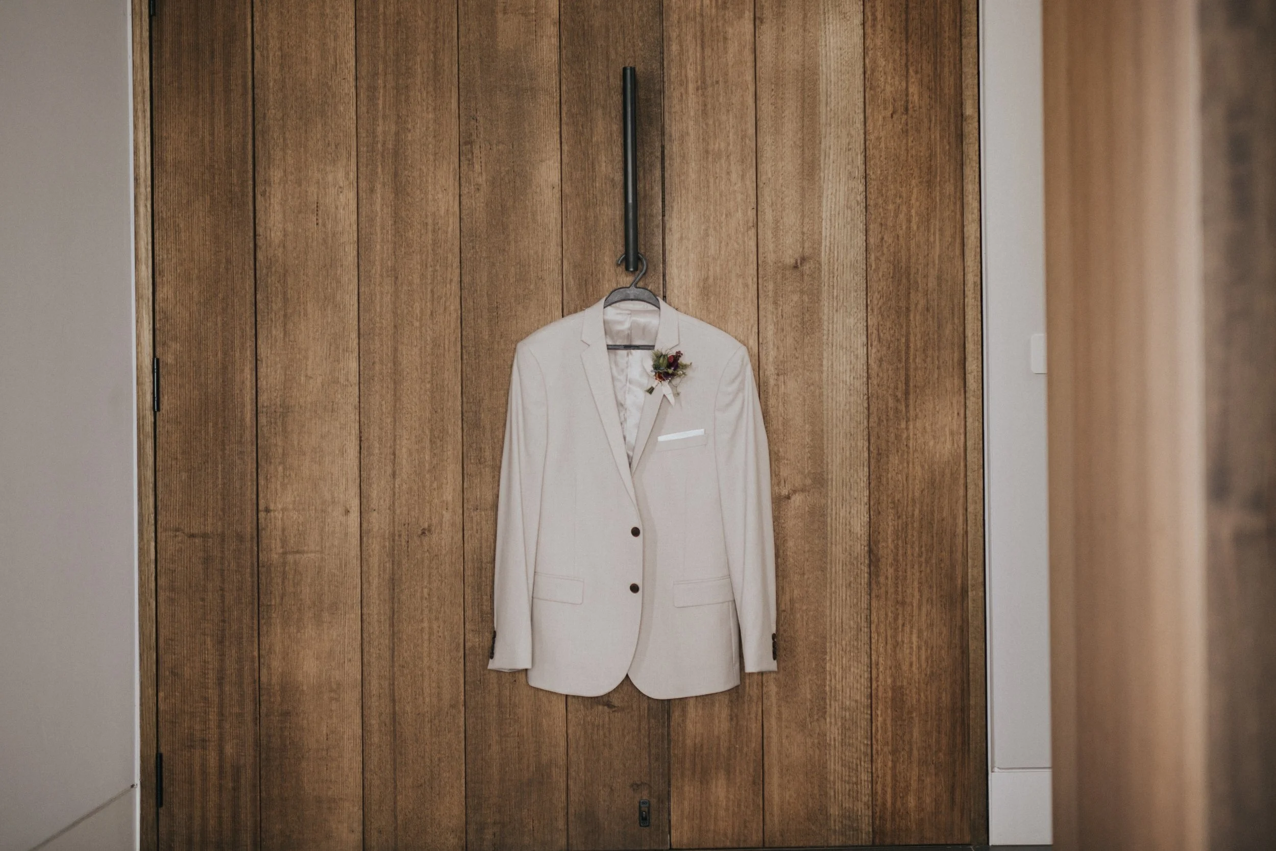 A white blazer hanging on a wooden wall with a boutonniere attached to the lapel.