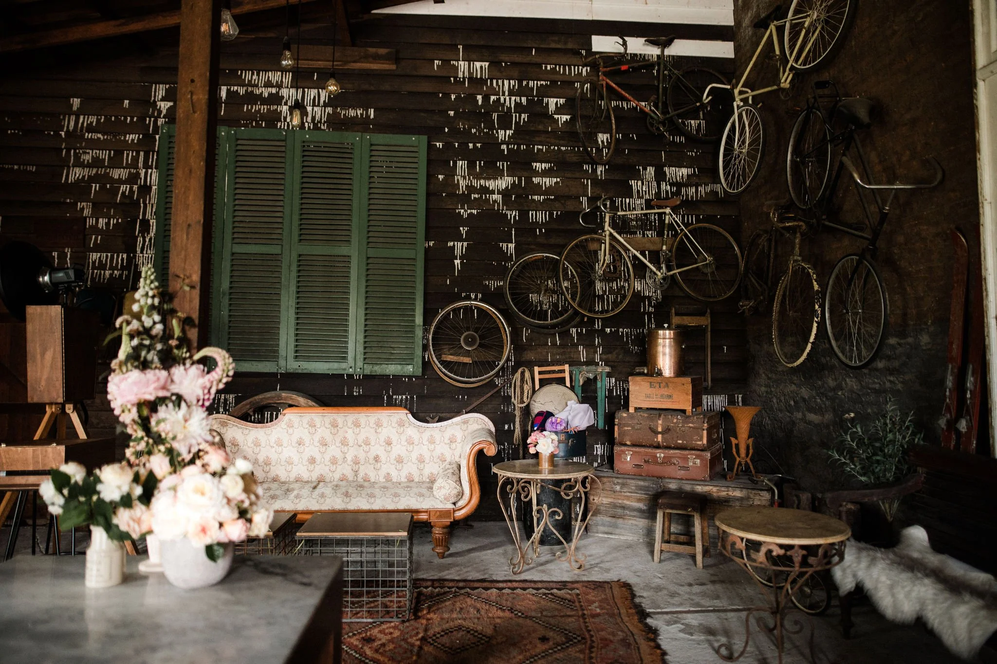 Interior of a rustic room with vintage bicycles hanging on the dark wooden wall, a green window shutter, an ornate sofa, a small round table with a floral bouquet, and various antique suitcases and decorative items.