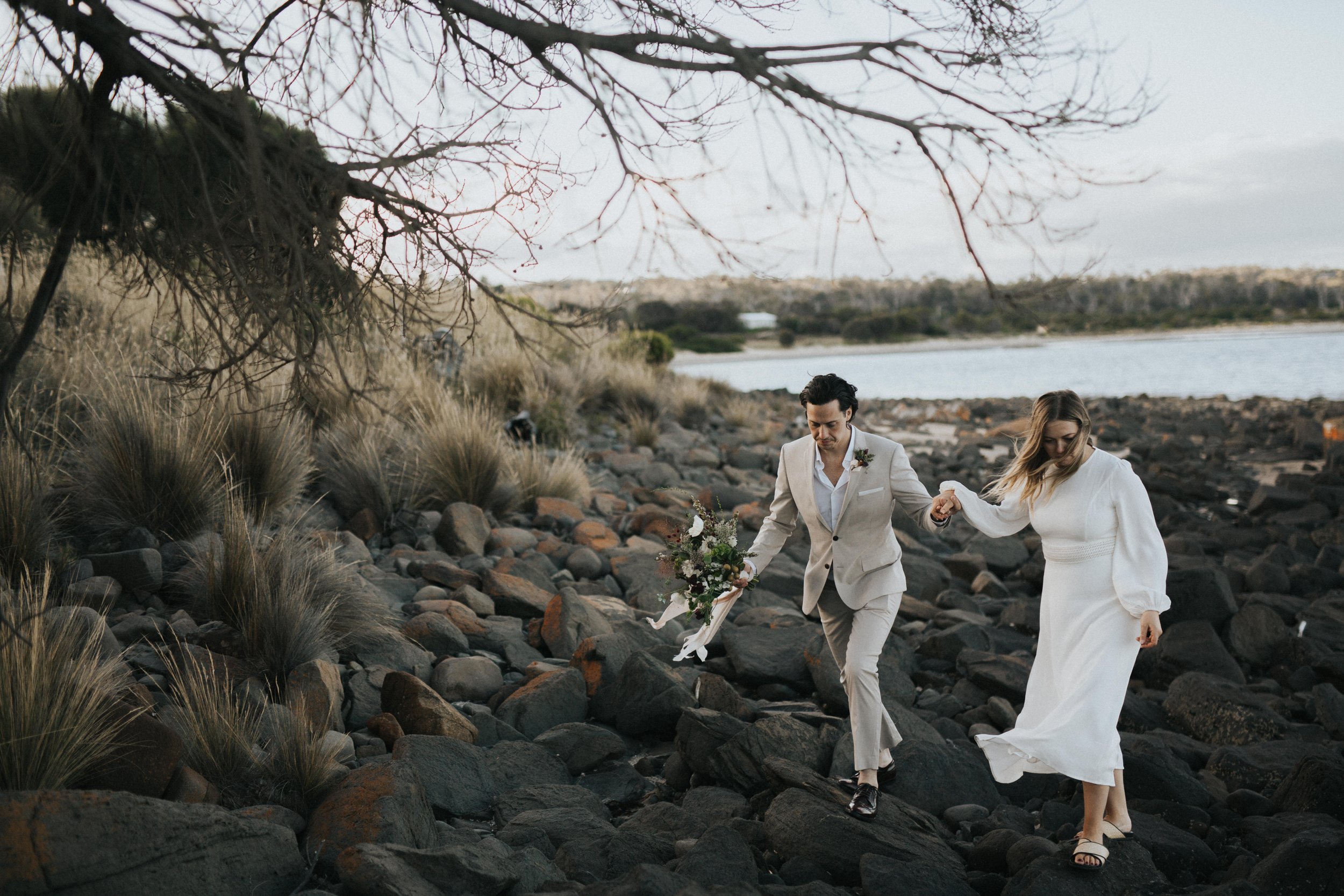 A couple in wedding attire walking on rocky terrain near a body of water, holding hands, with overhanging tree branches in the foreground.