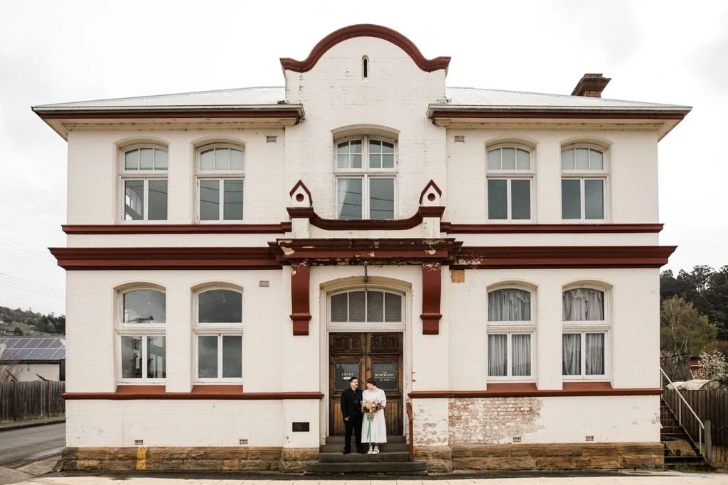 A two-story white house with red trim and a decorative front facade, with two people standing in front of the main door, one in dark clothing and the other in a white dress holding a bouquet.