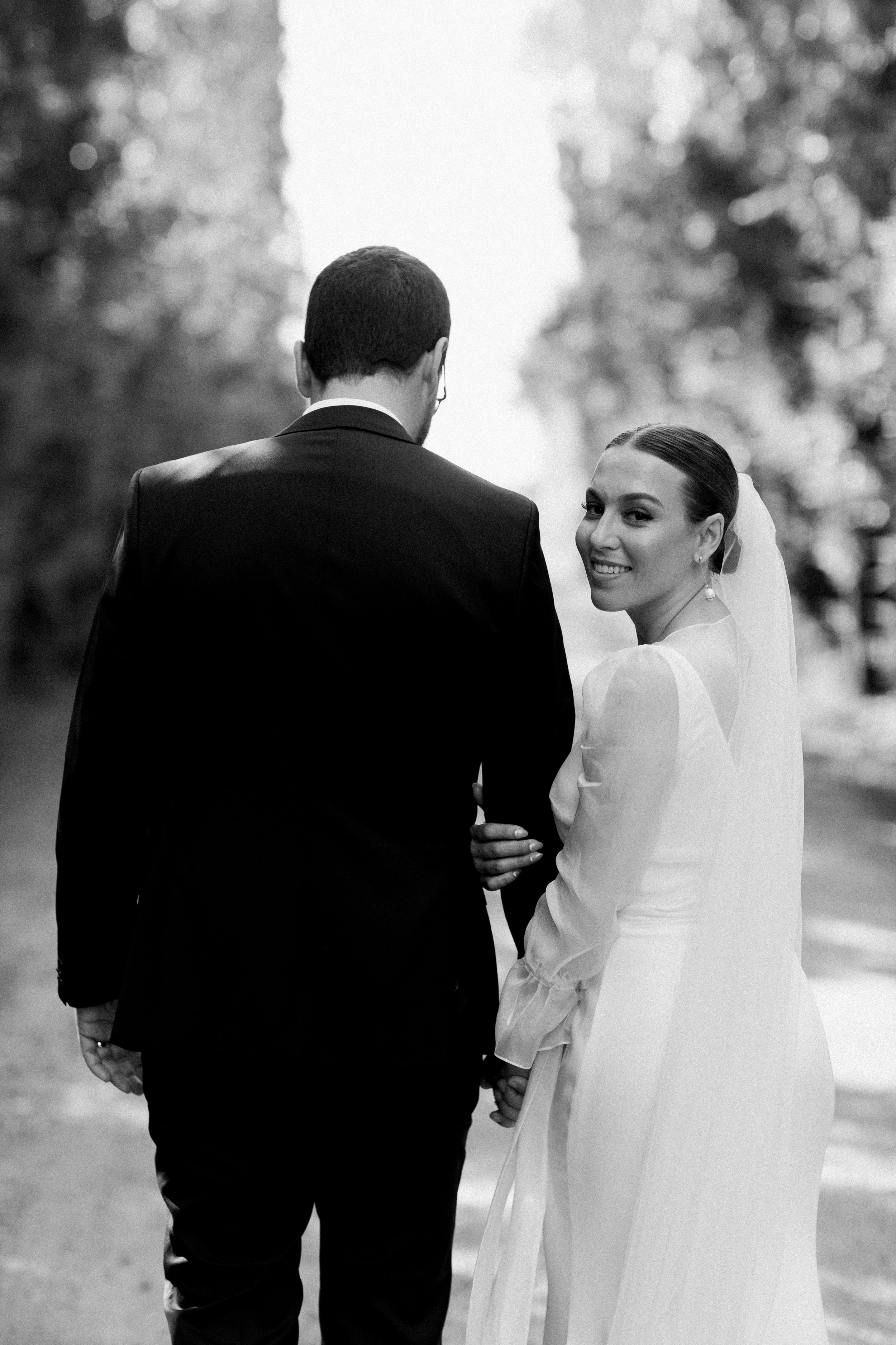 Black and white photo of a bride and groom holding hands, standing outdoors, with the bride smiling at the camera.