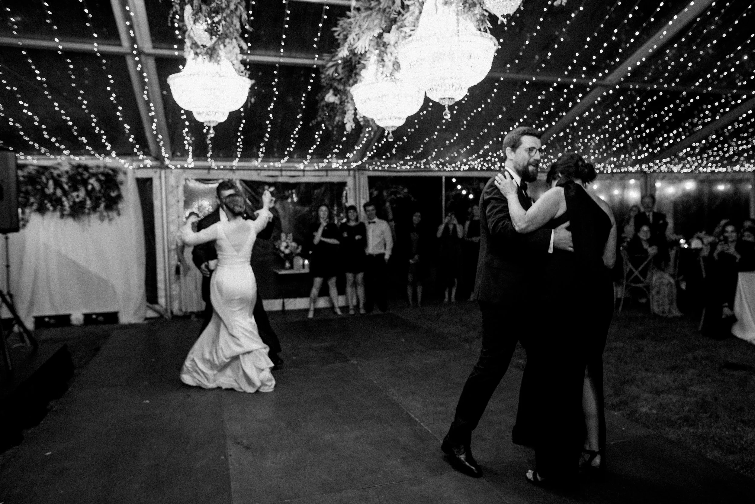 Couple dancing closely at a wedding reception, with the bride and groom in the background dancing and guests watching under fairy lights and chandeliers.