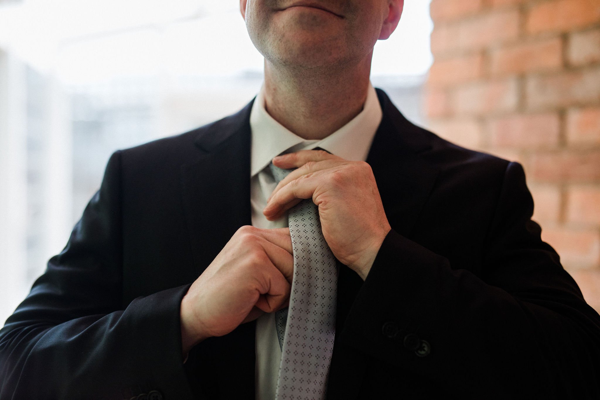 A man in a black suit adjusting his grey tie in front of a brick wall and window.