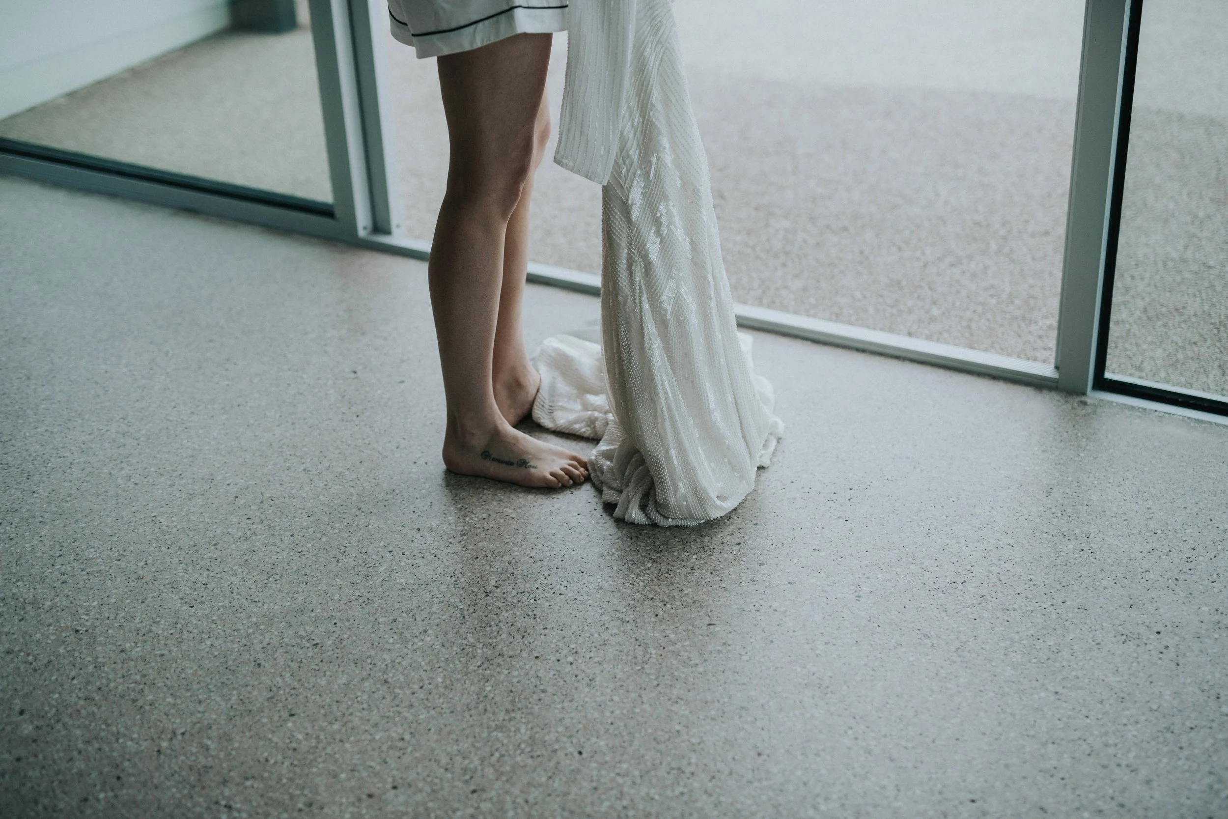 Person standing barefoot on a terrazzo floor near a large window, holding a white, textured fabric or dress.