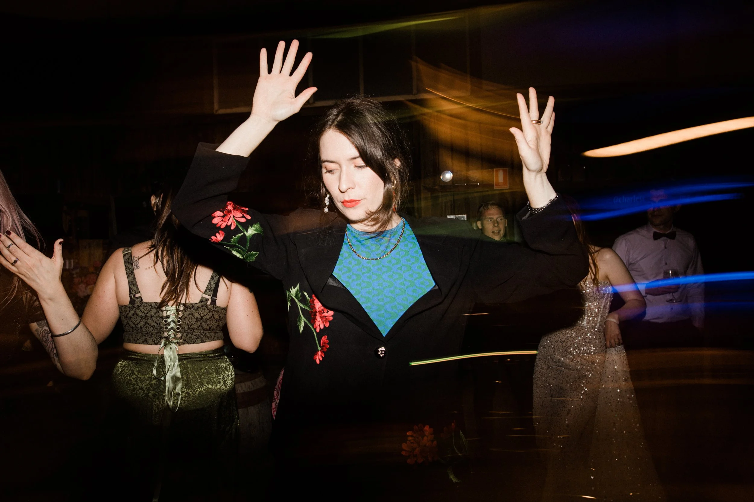 A woman with dark hair and red lipstick dancing with eyes closed, wearing a black blazer with floral embroidery and a blue patterned top, at a party with other people in elegant attire.