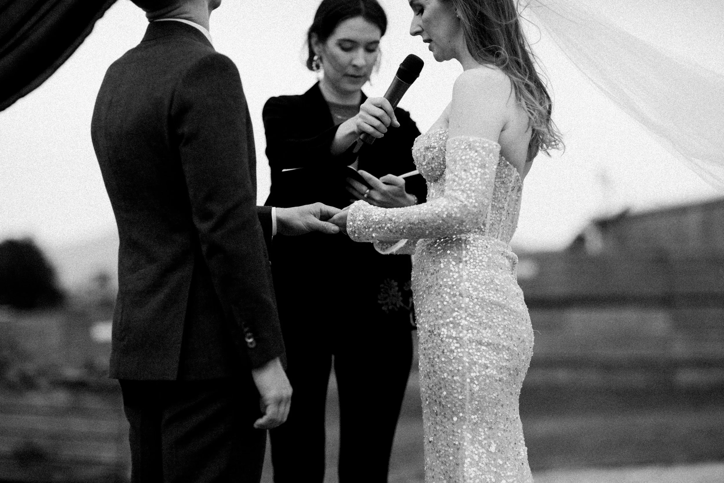 Black and white photo of a wedding ceremony with a bride and groom holding hands, an officiant reading from a book, and a woman holding a microphone.