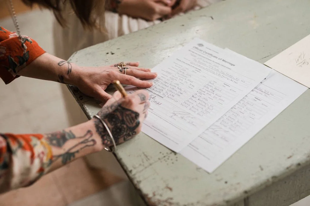 Person signing a document on a worn desk, with tattoos visible on their arm and hand.