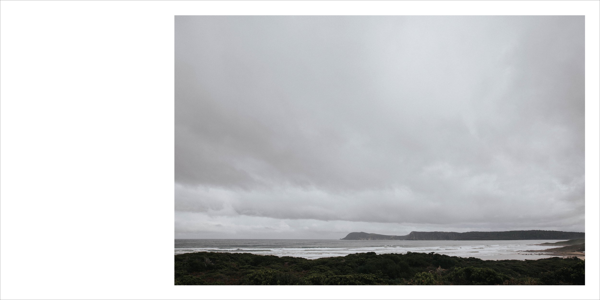 Overcast sky over a coastal landscape with a beach, ocean waves, and a distant headland covered with vegetation.