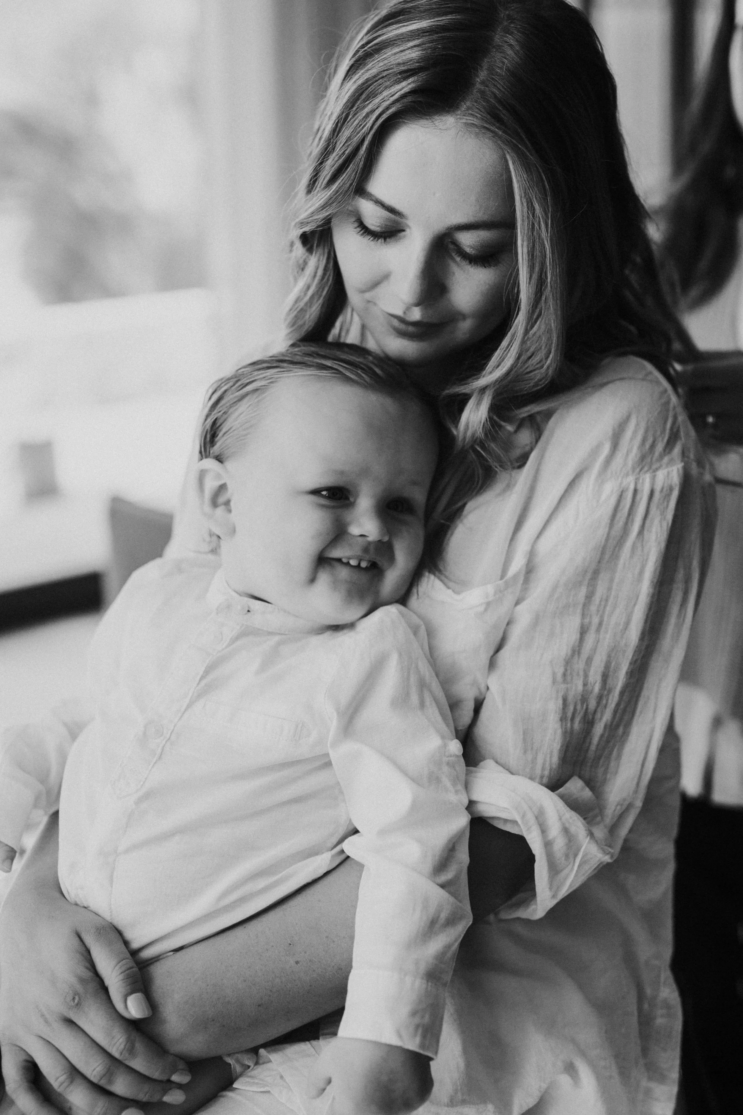 A woman holding a smiling young boy in a cozy indoor setting, both dressed in light-colored clothing.
