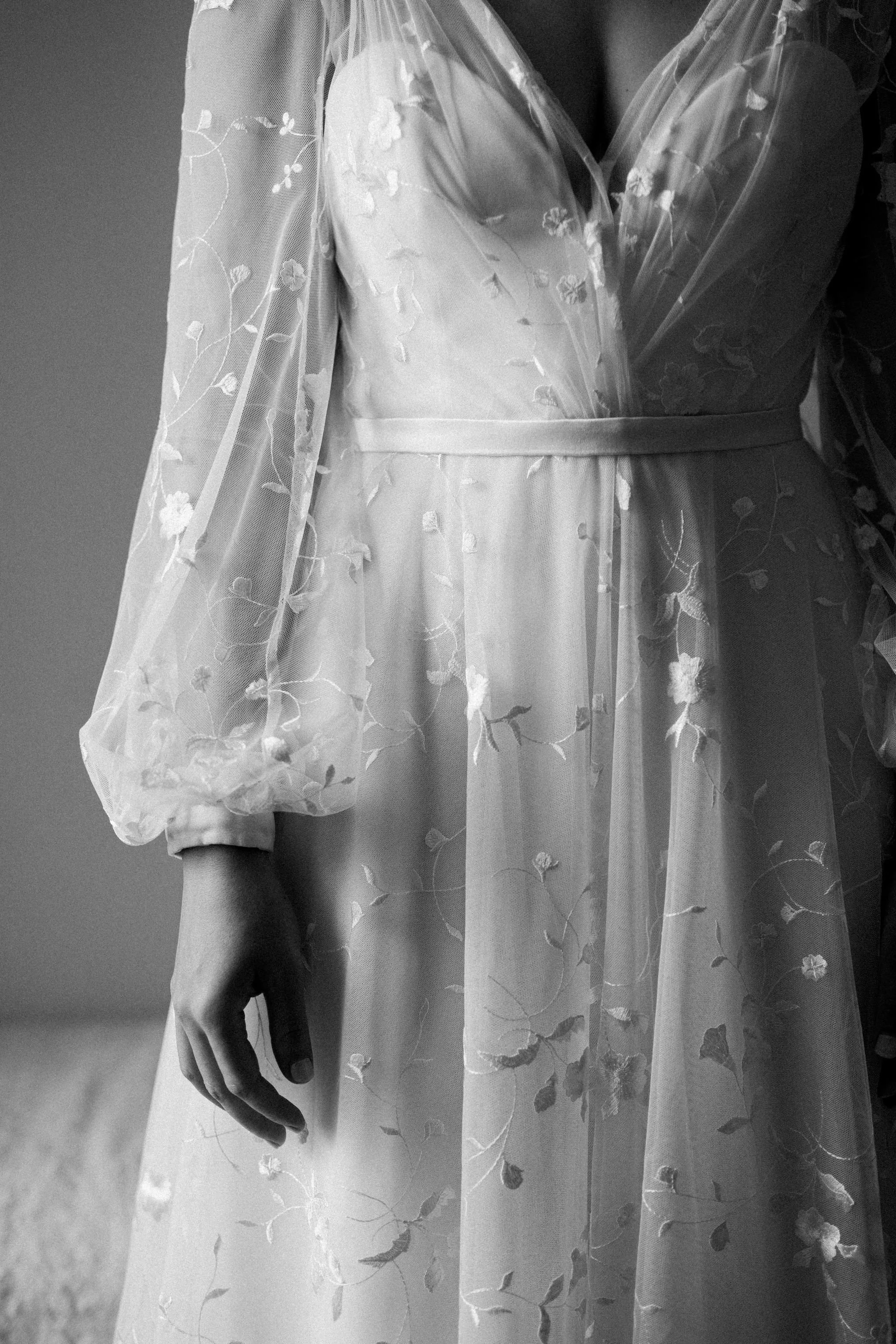 Close-up of a person wearing a delicate, embroidered sheer dress with floral patterns. The image is in black and white, emphasizing the fabric's intricate details.