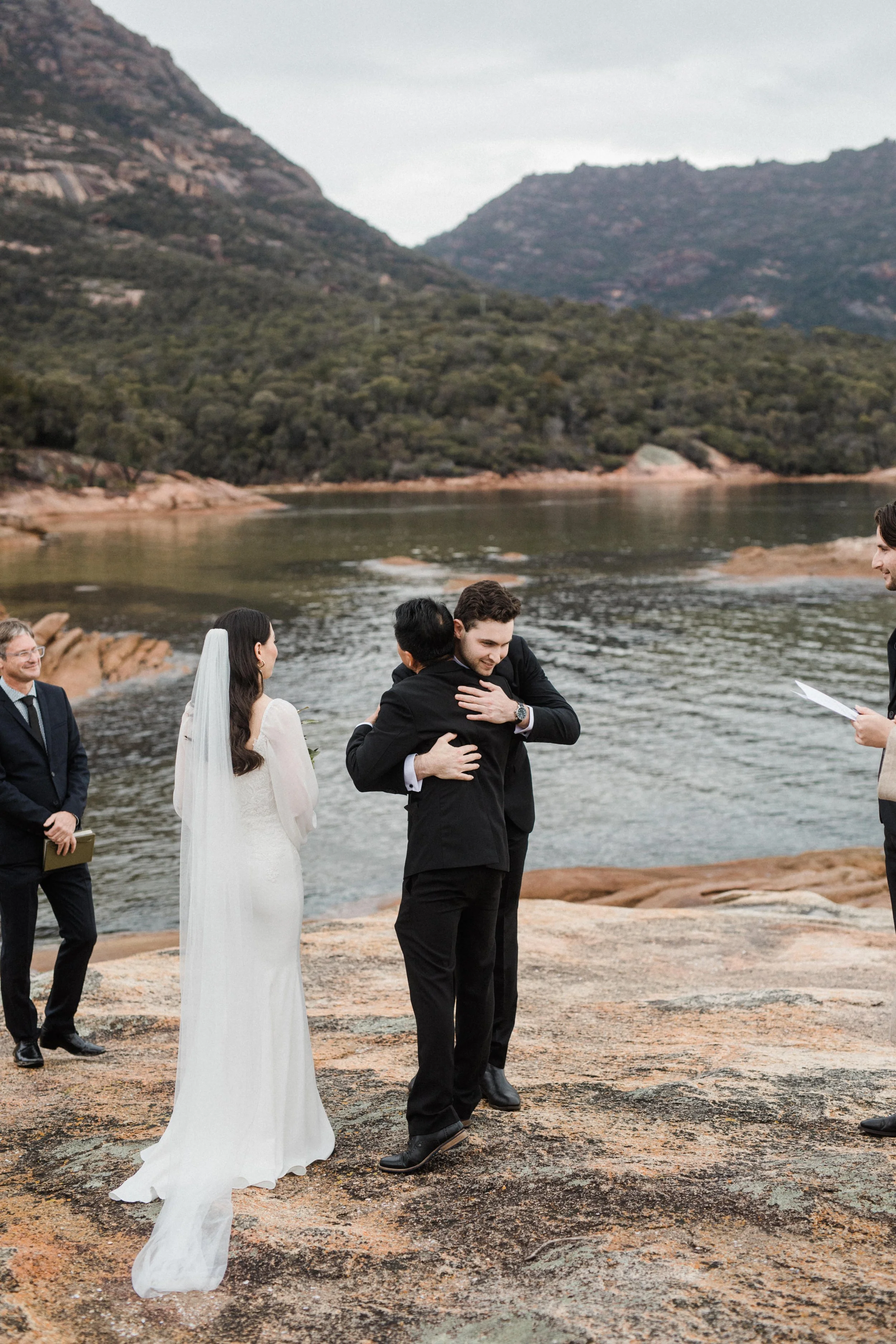A wedding ceremony takes place outdoors by a river, with mountains and trees in the background. The bride and groom are hugging, while an officiant reads from a paper. Two other men, dressed in suits, are standing nearby.
