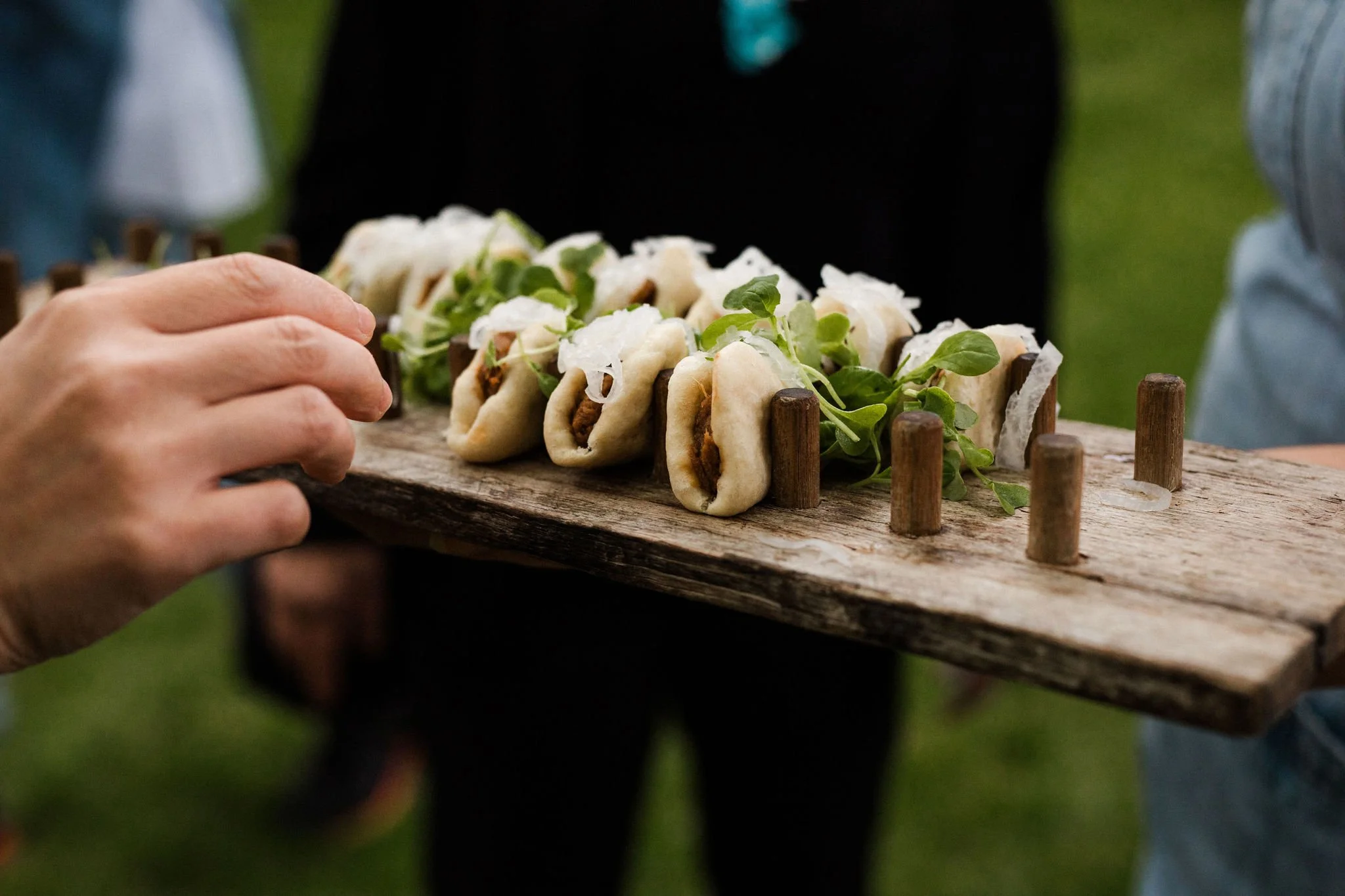 Hand reaching to pick up a small sandwich on a rustic wooden serving board, with several similar sandwiches decorated with greens.