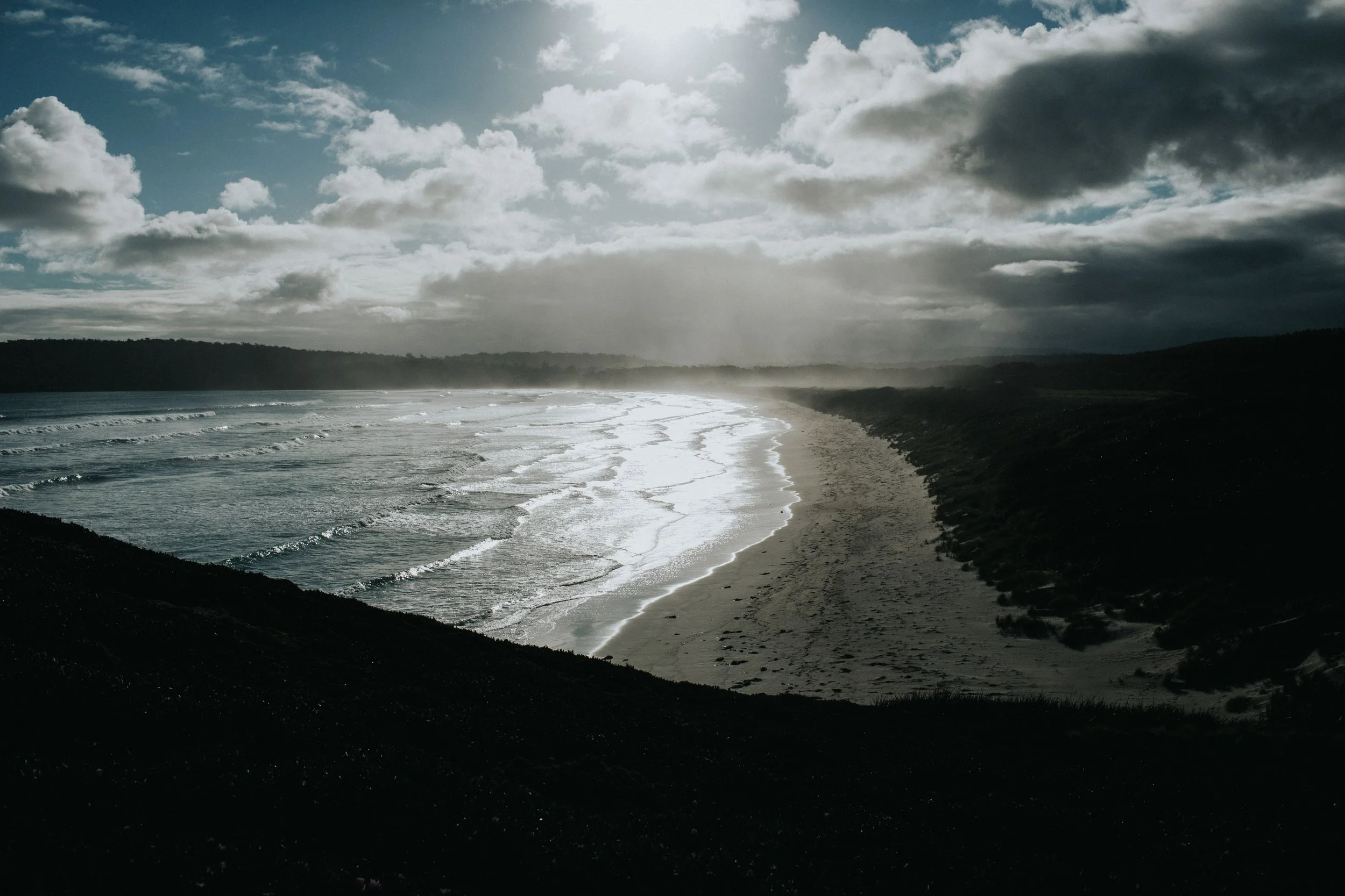 A beach with dark sand, rolling waves, and cloudy skies with sunlight breaking through.