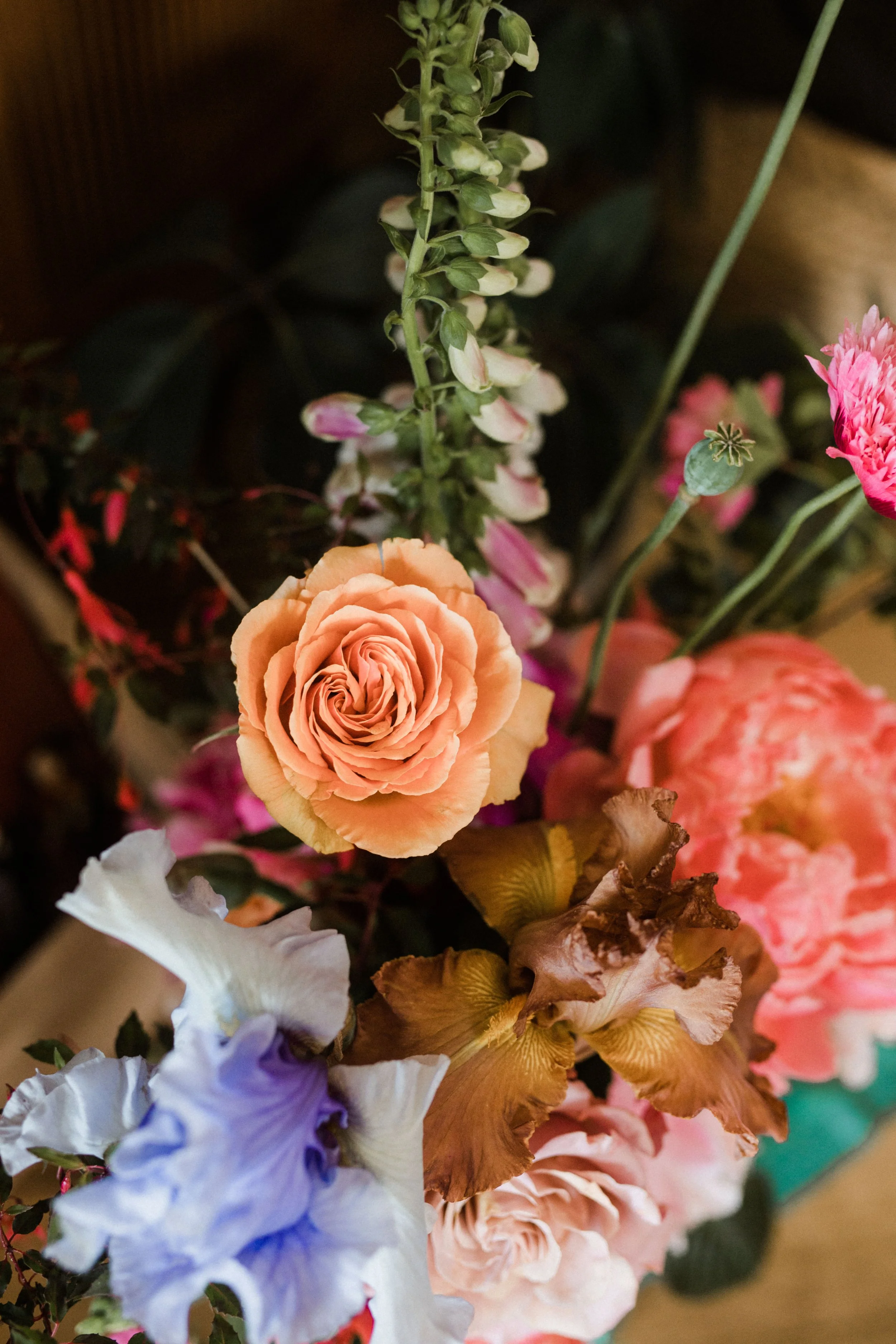 Close-up of a colorful bouquet with various flowers, including a peach-colored rose, purple and white irises, pink and white carnations, and green foliage.