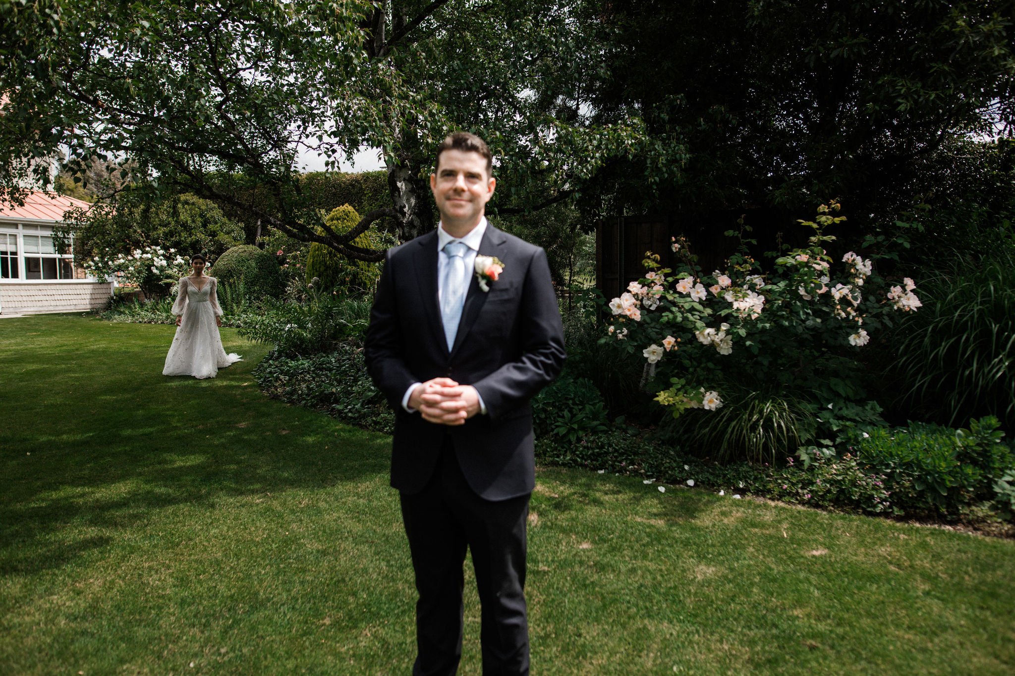 A groom in a suit with a boutonniere stands in the foreground, smiling, while a bride in a wedding dress walks toward him in a lush garden.