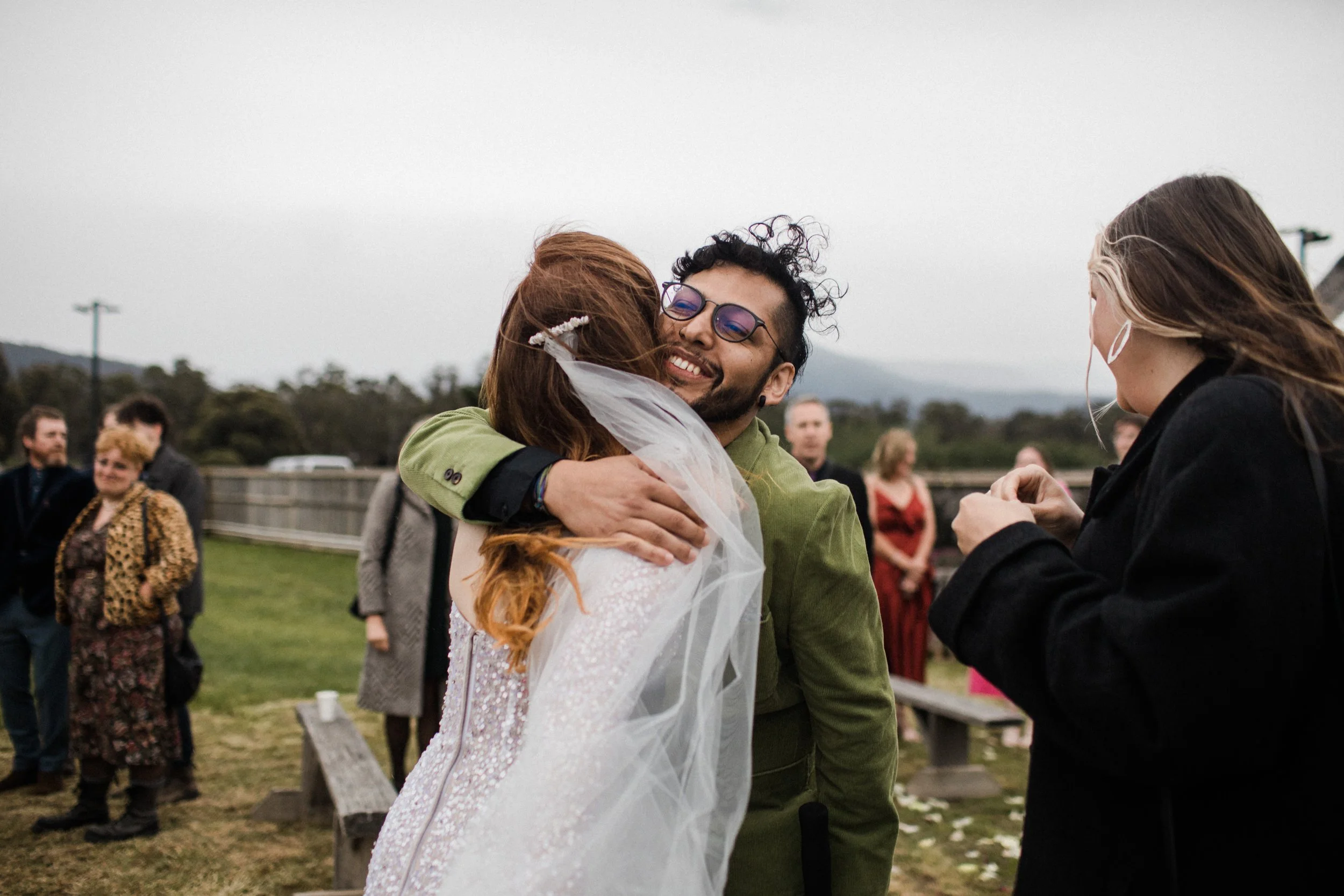 A man and woman hugging at an outdoor wedding, other guests in the background on a cloudy day.
