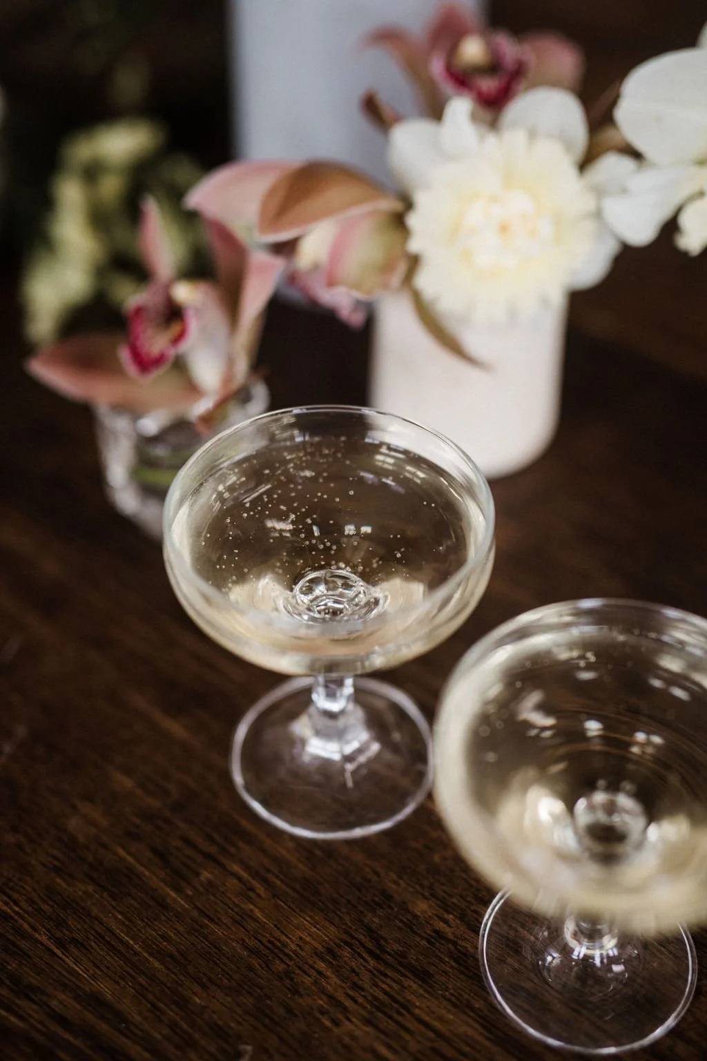 Two champagne glasses filled with sparkling wine on a wooden table with flower arrangements in the background.