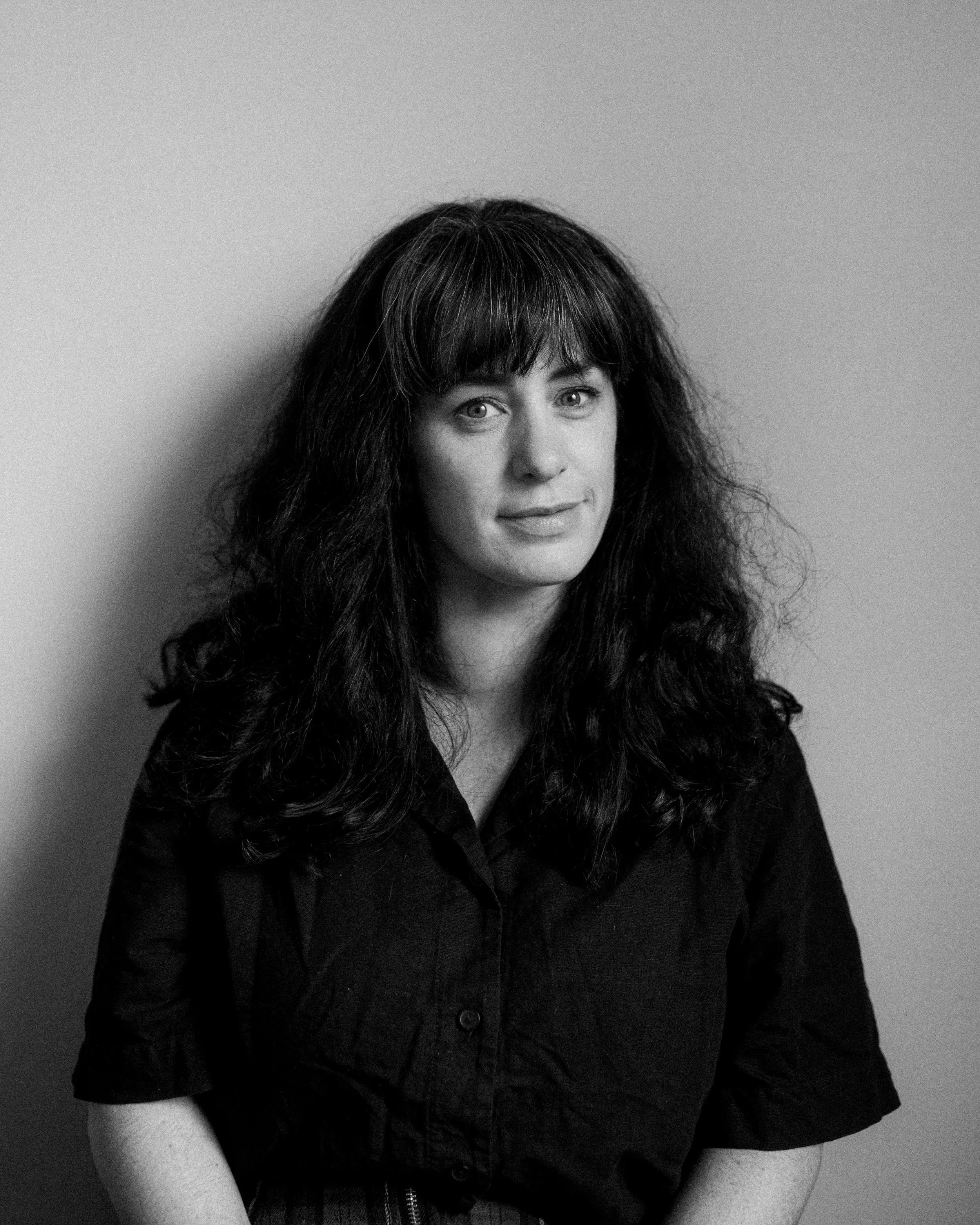Black and white portrait of a woman with dark, curly hair and bangs, wearing a dark blouse, against a plain background.