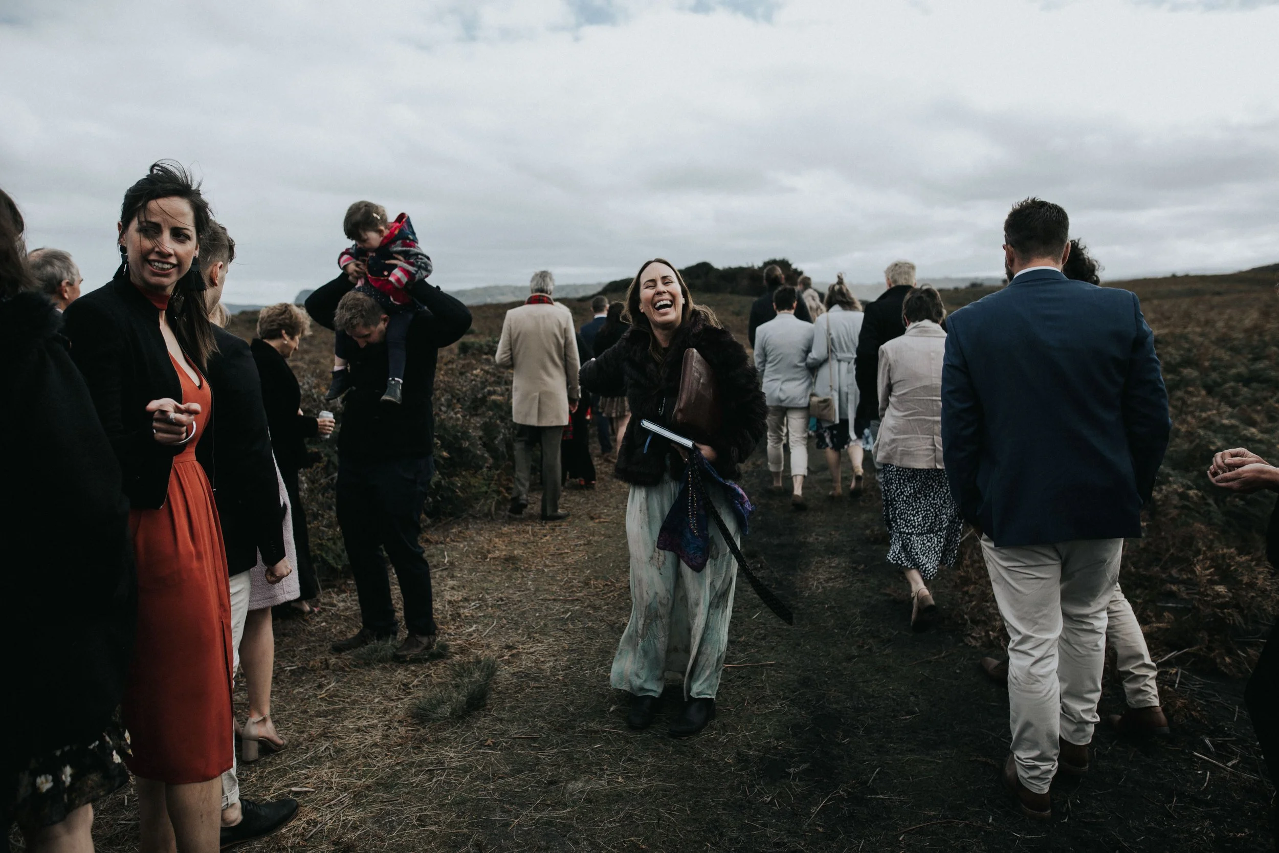 People walking outdoors on a cloudy day, dressed in formal and semi-formal clothing, some smiling and talking, with a woman in a fur coat laughing at the center.
